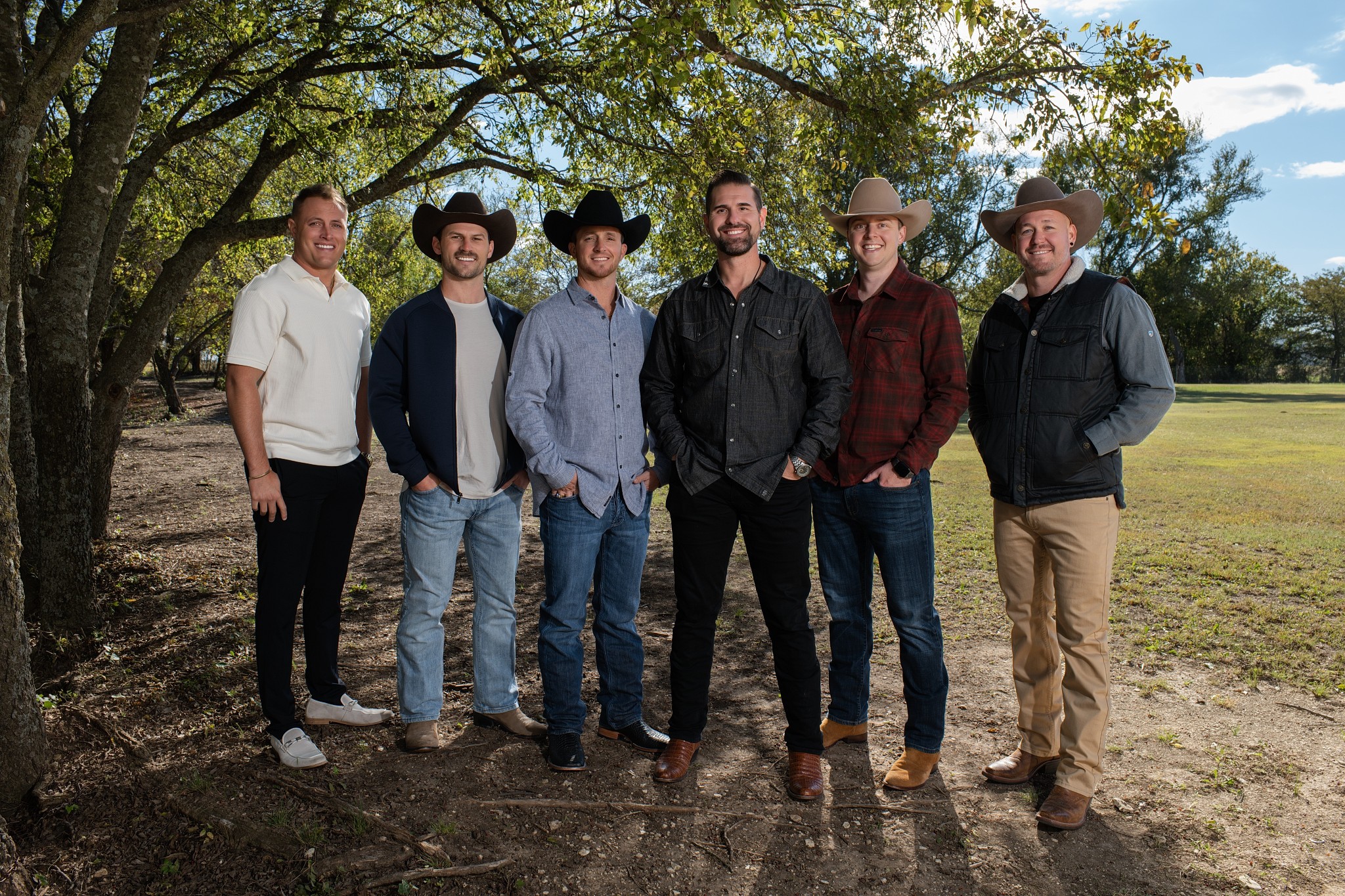 Group of six men standing outdoors under trees, dressed casually, with a grassy field in the background.