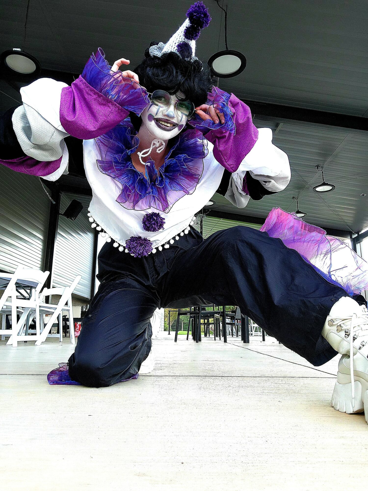 Person dressed as a clown with colorful costume, makeup, and a hat, crouching outdoors with a playful pose.