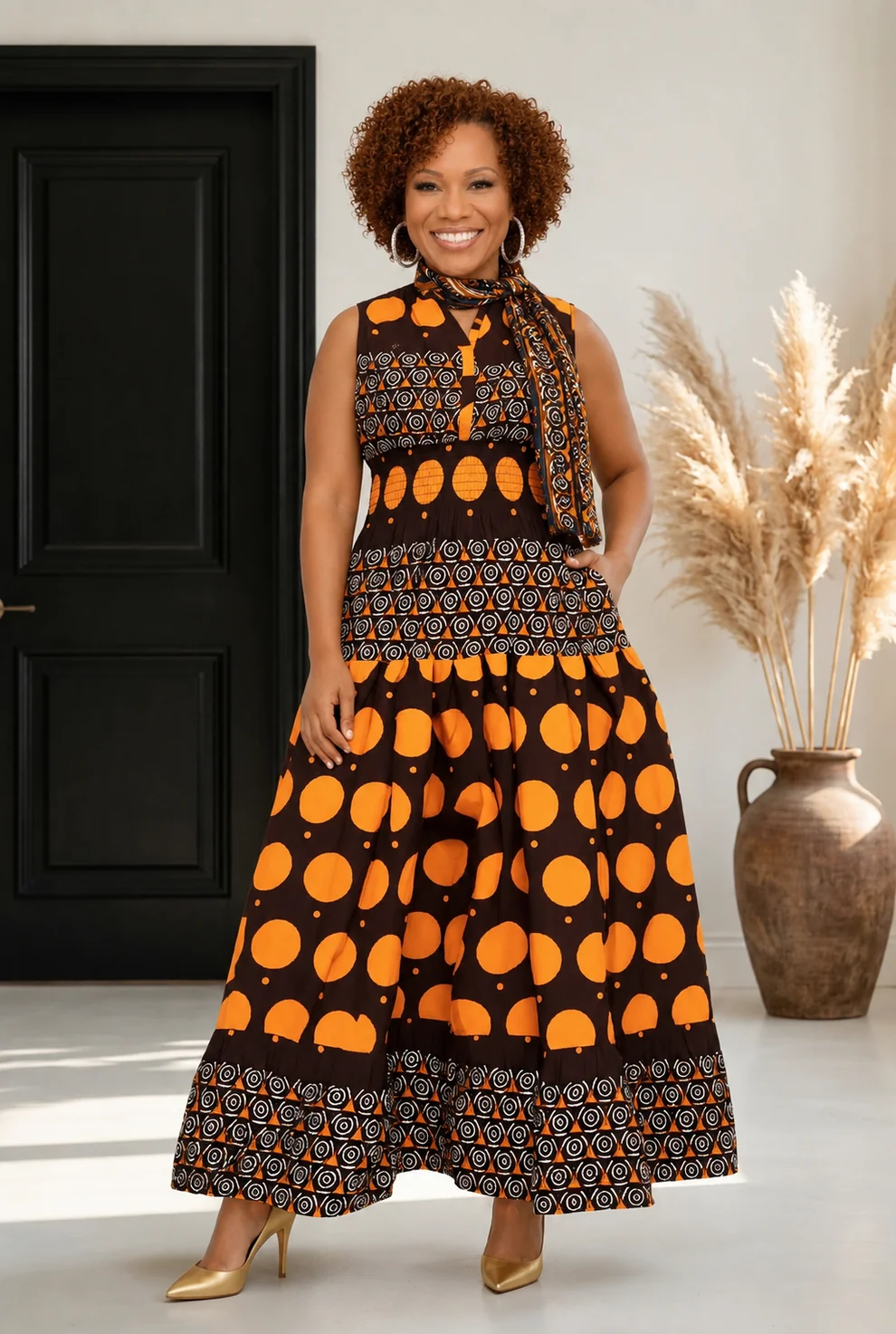 Smiling woman in a black and orange patterned dress standing indoors with a black door and a large vase of pampas grass behind her.
