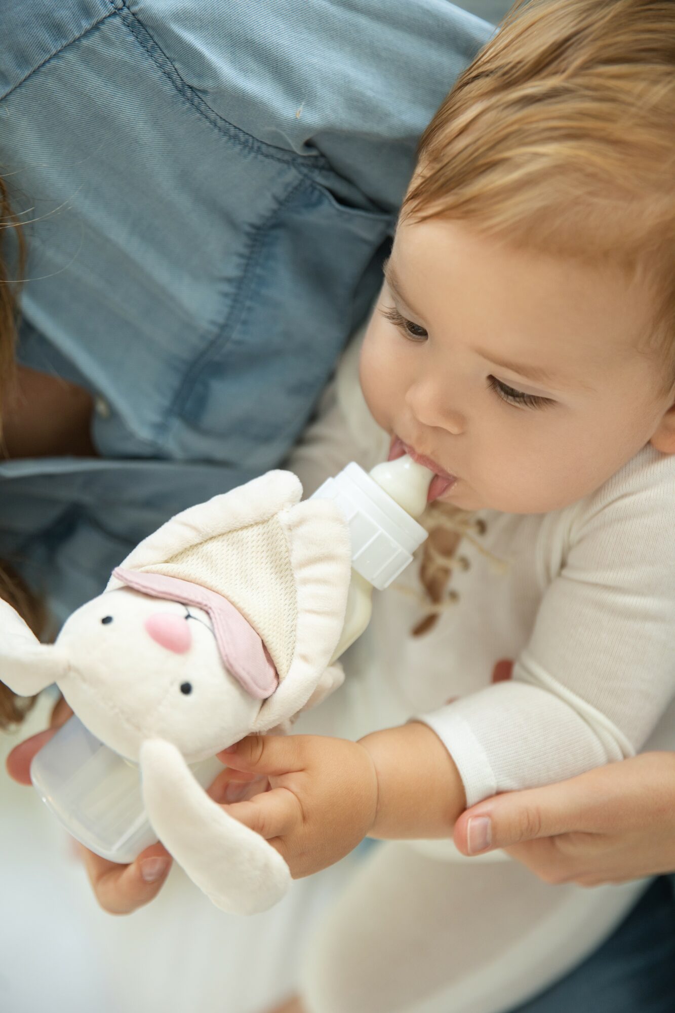Young child with light hair drinks from a bottle held by an adult, holding a plush toy, in a close-up shot.