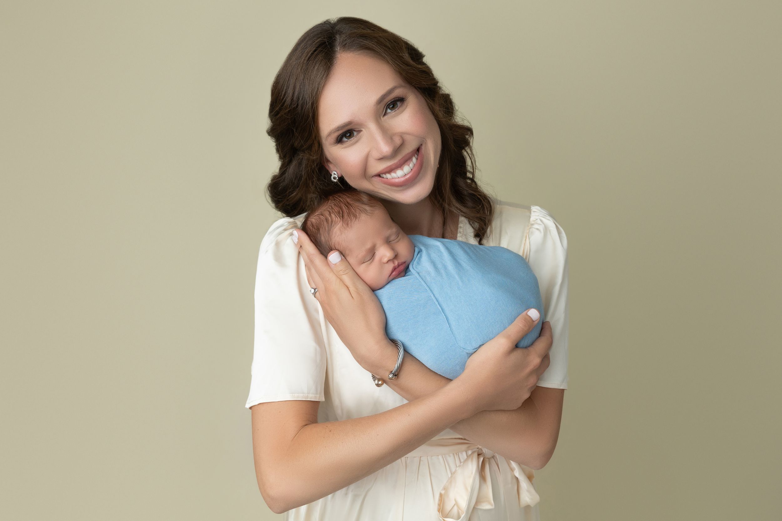 Woman holding a sleeping baby wrapped in a blue blanket, smiling, against a plain background.