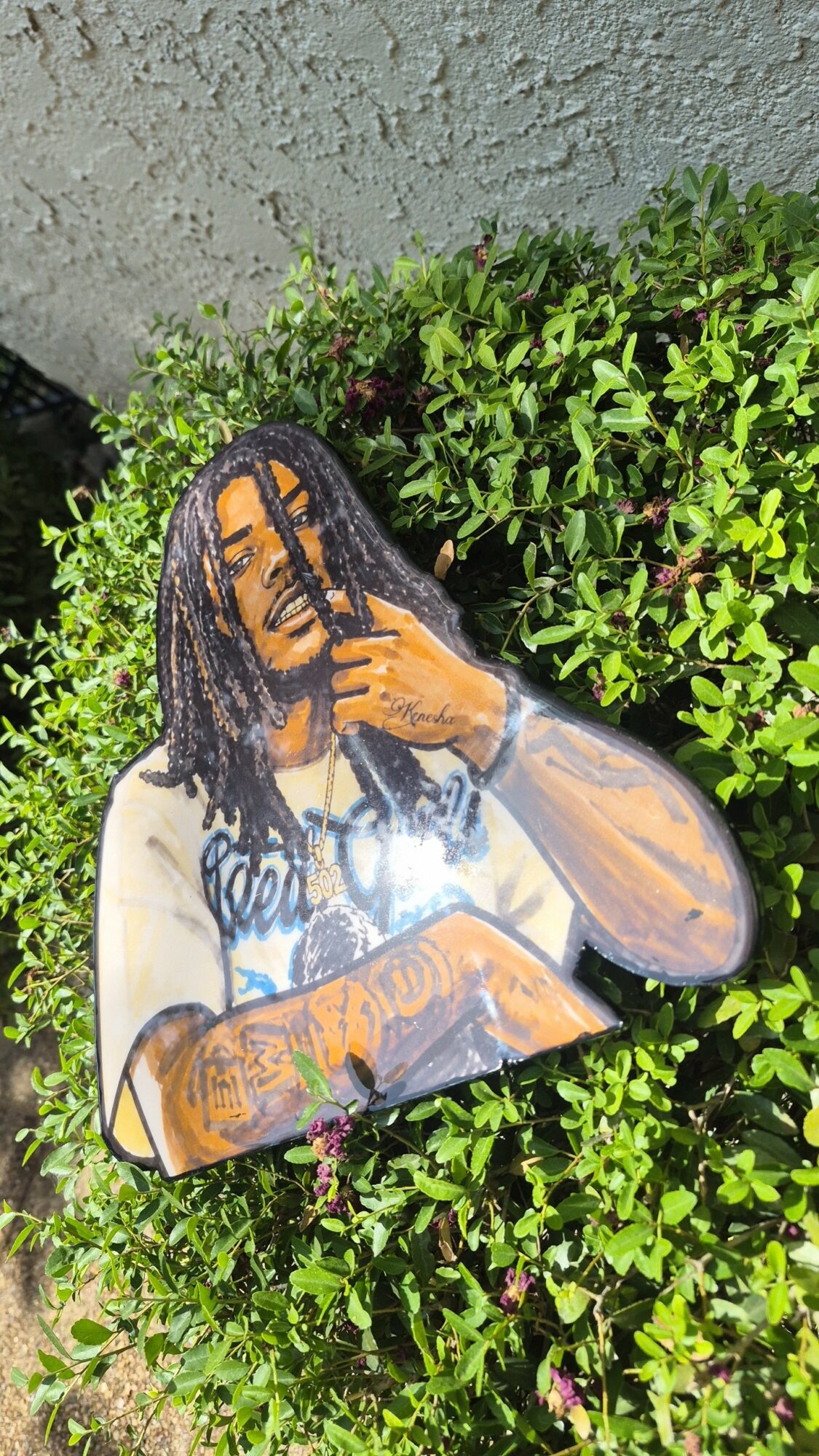 A painted cutout of a person with dreadlocks, holding a book and making a peace sign, placed outdoors near green bushes.
