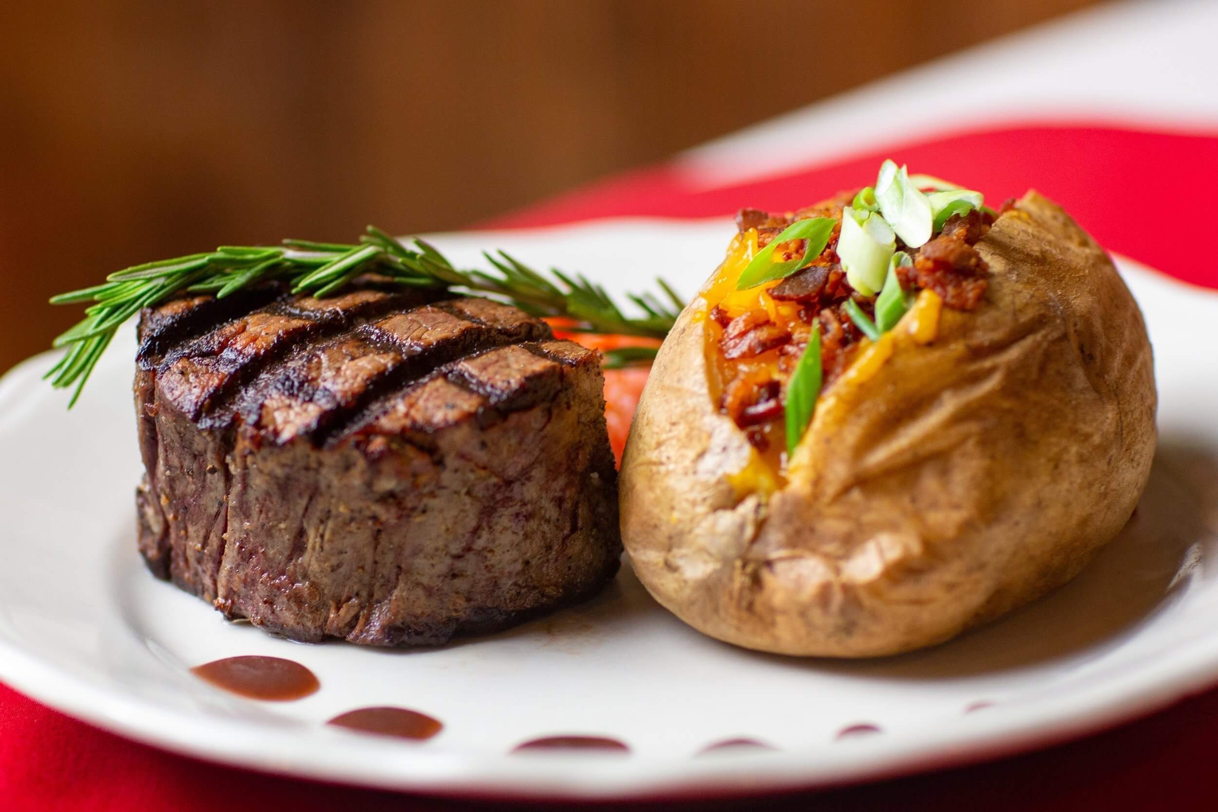 Grilled steak with grill marks, garnished with rosemary, served with a baked potato topped with vegetables and green onions.