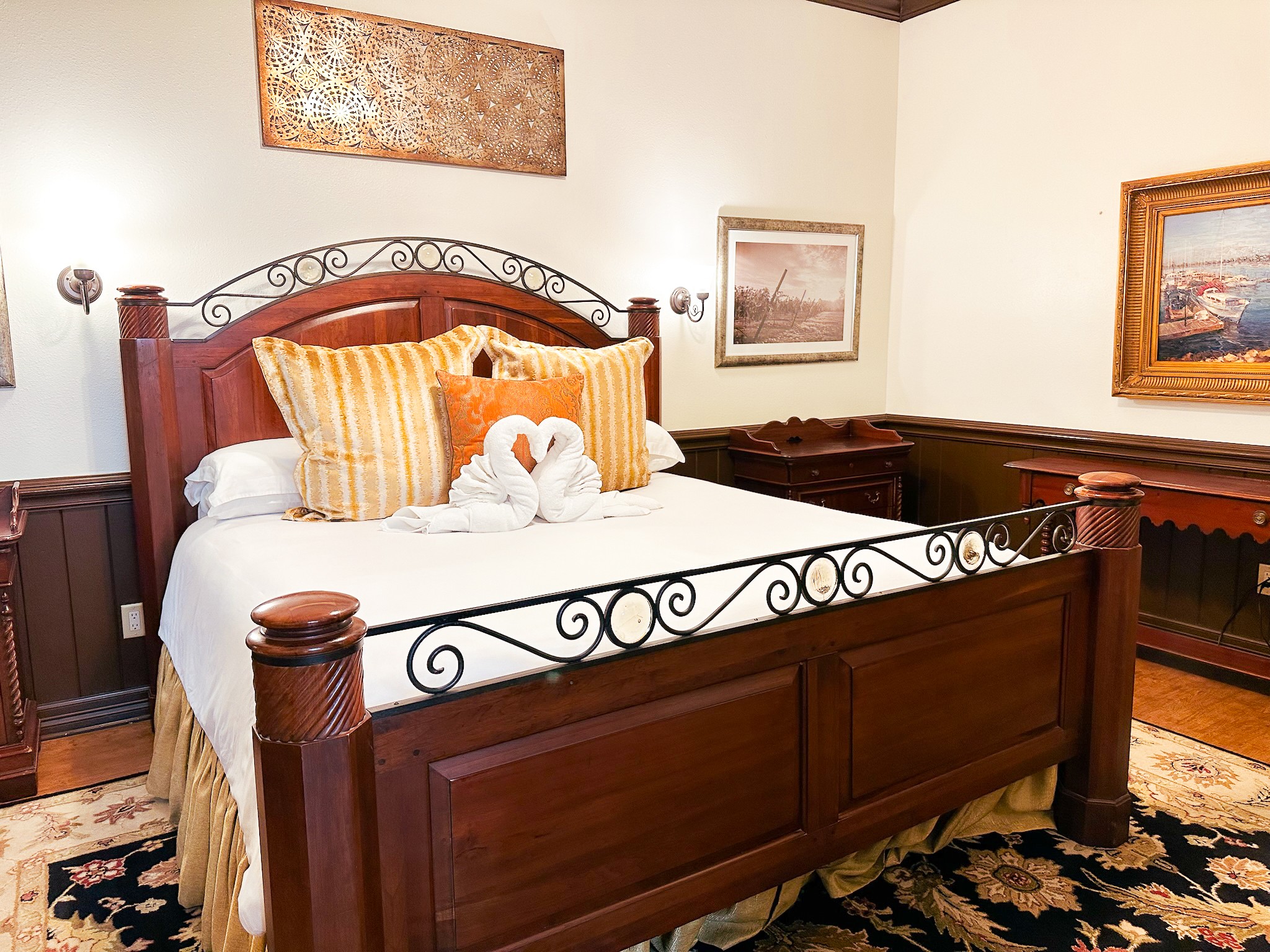 A bed with a wooden frame and decorative metal headboard, two swan towels, and pillows, in a room with artwork and a rug.