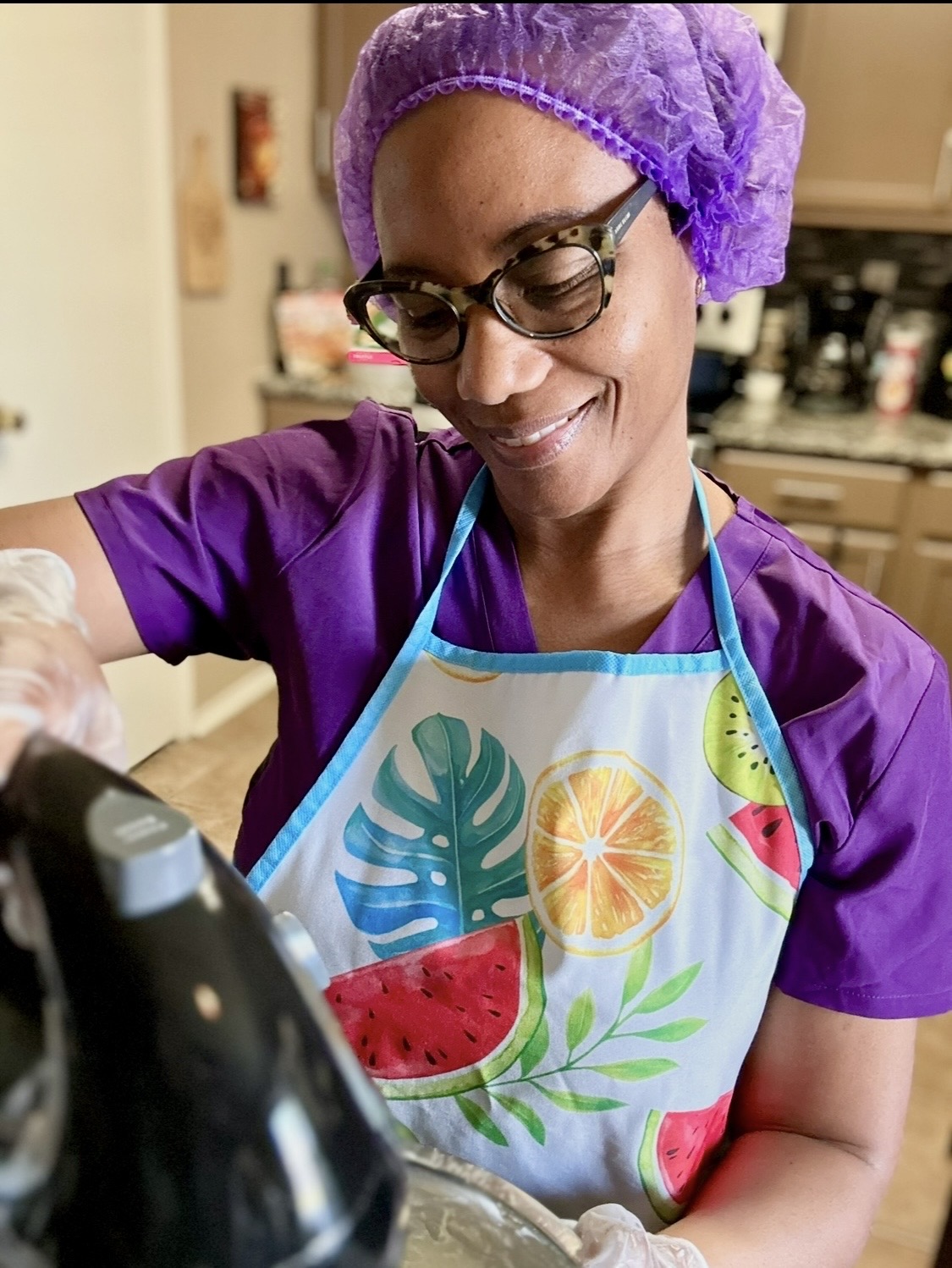 Person wearing purple scrubs, glasses, and a purple hair cover, smiling while cooking in a kitchen, wearing a colorful apron with fruit and leaf patterns.
