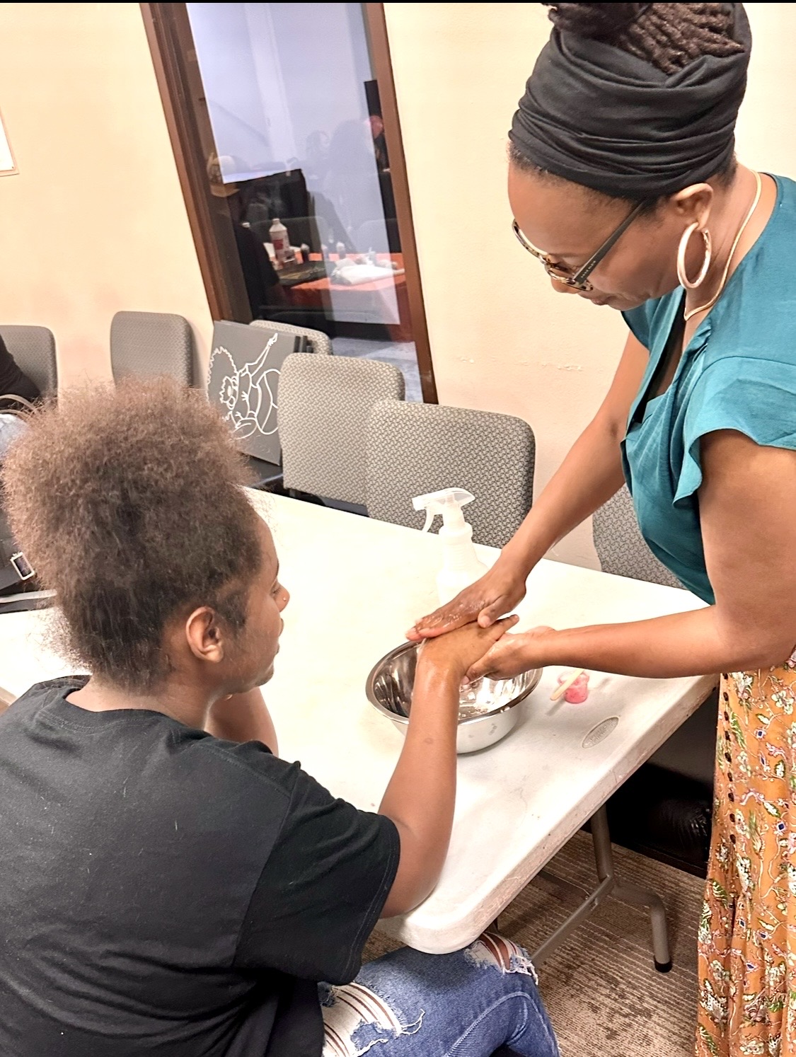 Woman with glasses and headscarf helping a child with hair at a table in a room with chairs and a window.