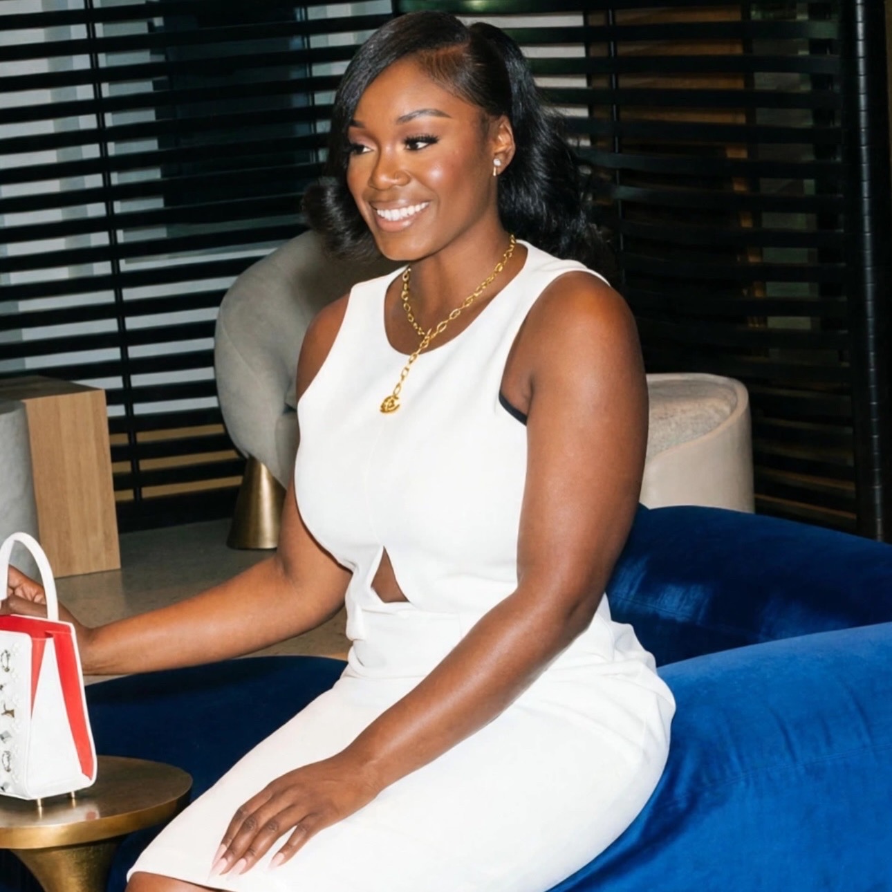 Smiling woman in white dress sitting on a blue couch, with black blinds in the background.