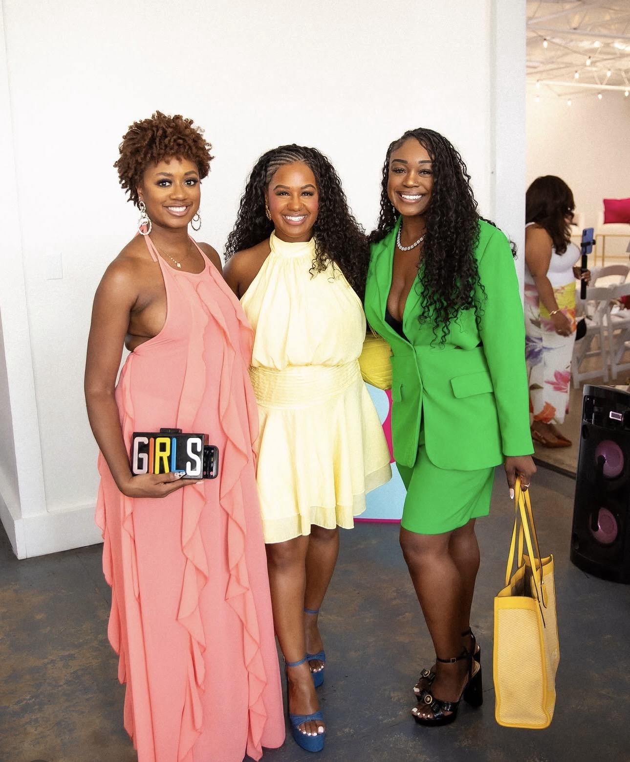Three women in colorful dresses smiling, standing together indoors, with a white wall background and other people in the distance.