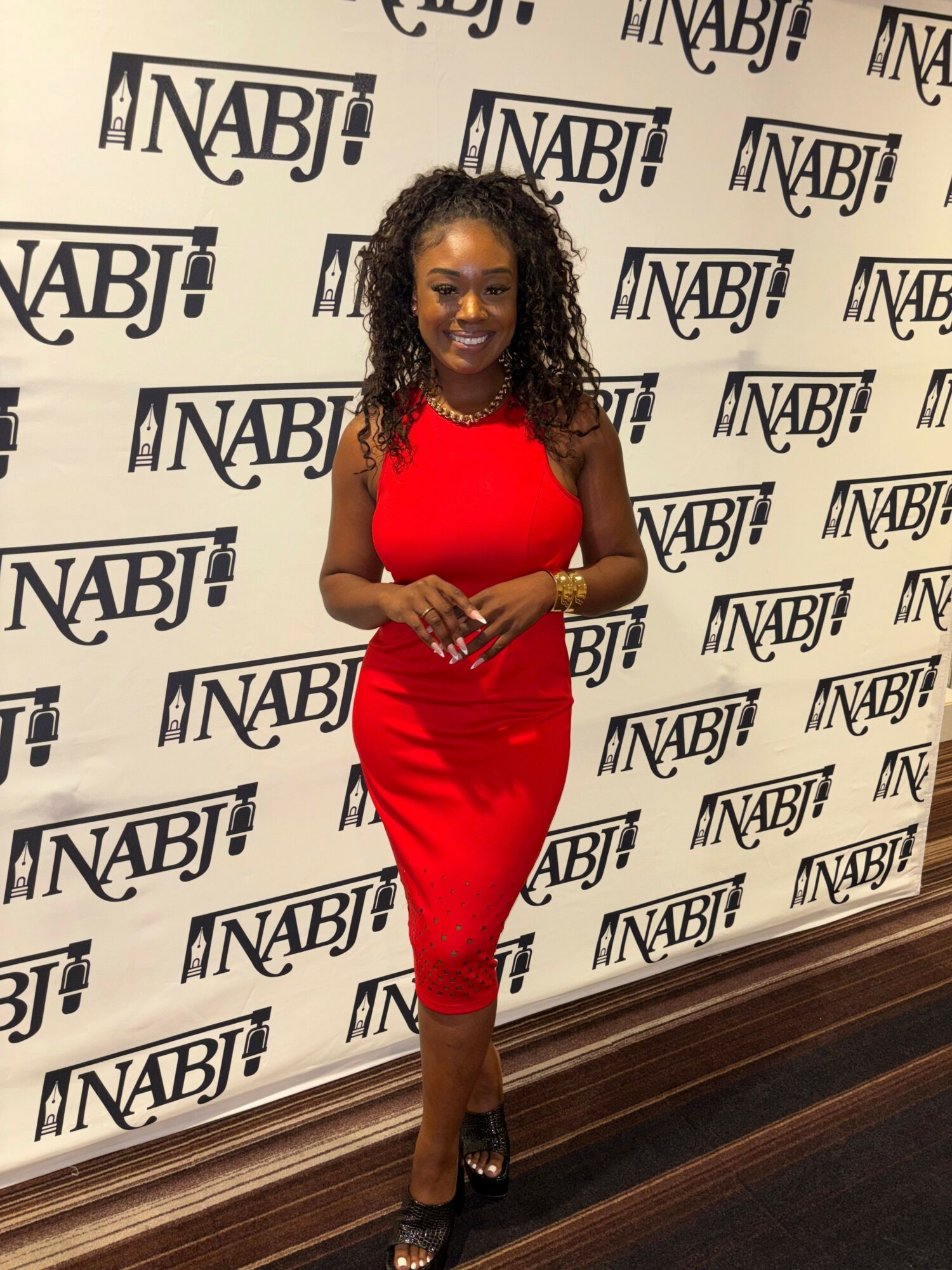Woman in red dress standing on a patterned floor with NABJ logo backdrop, holding a phone, smiling.