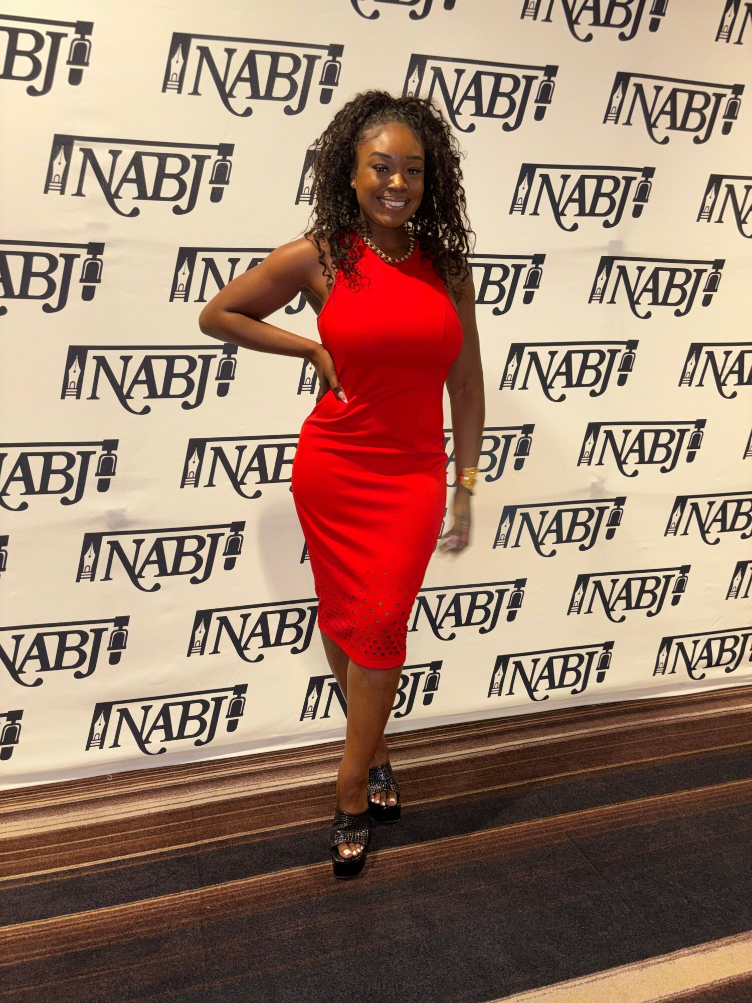 Woman in red dress standing in front of NABJ backdrop, posing with hand on hip, smiling, with dark hair and heels.