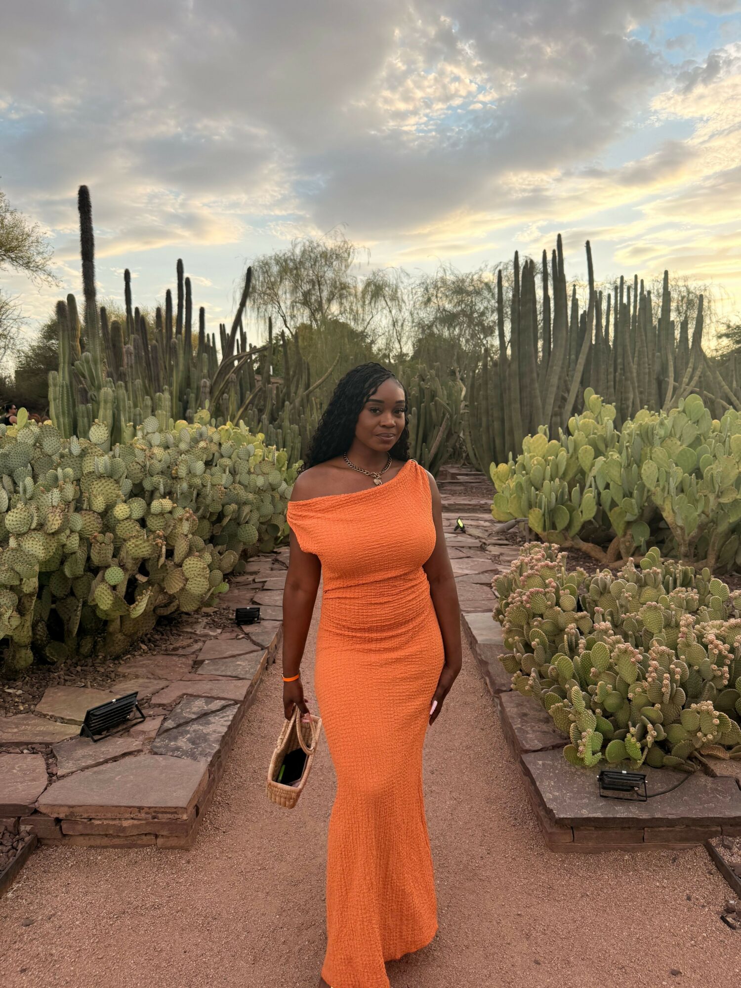 Woman in orange dress walking along a cactus garden path during sunset, holding a bag, with cloudy sky overhead.