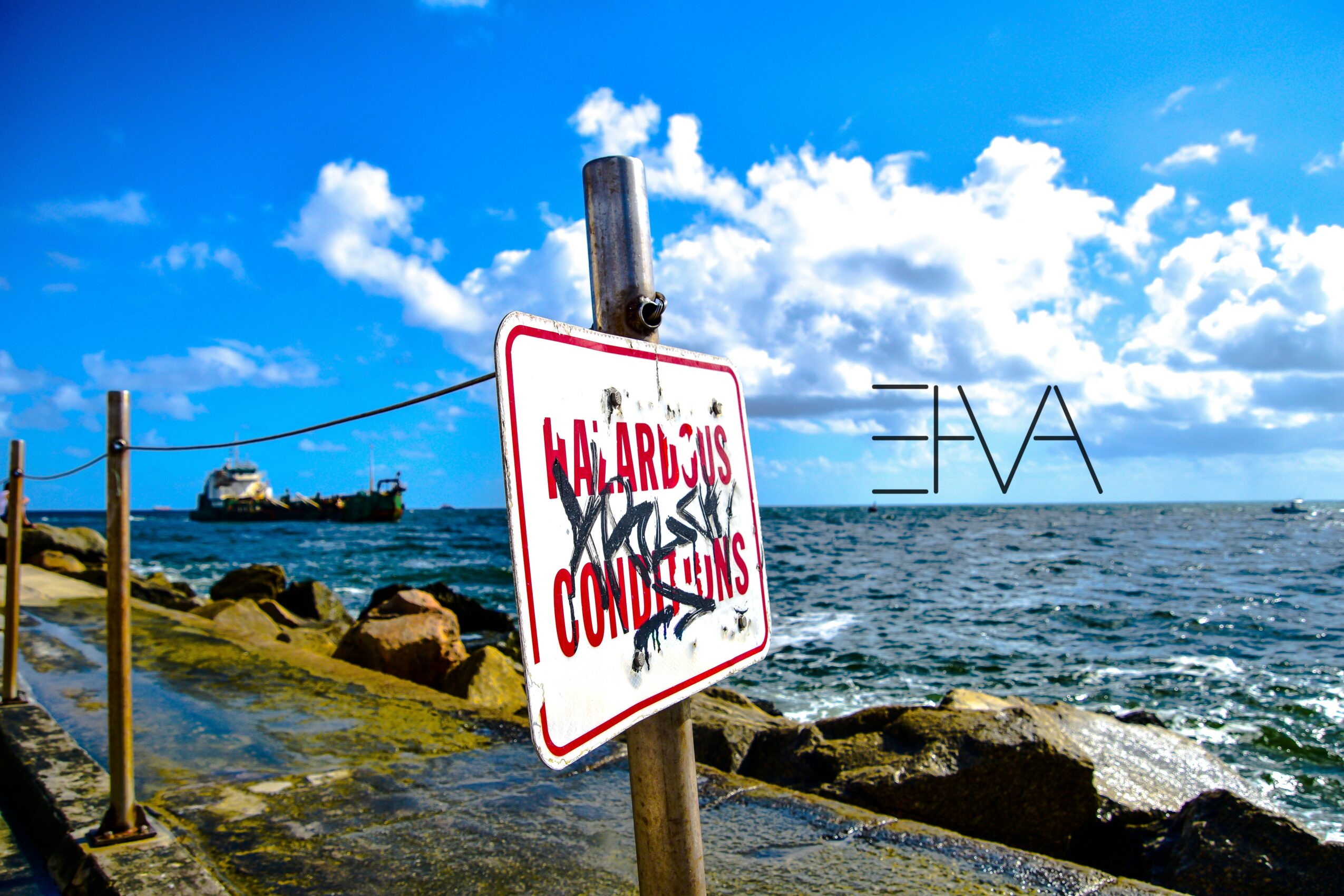 Sign on a pole near rocks by the sea with a boat and a large metal structure in the background, under a blue sky.