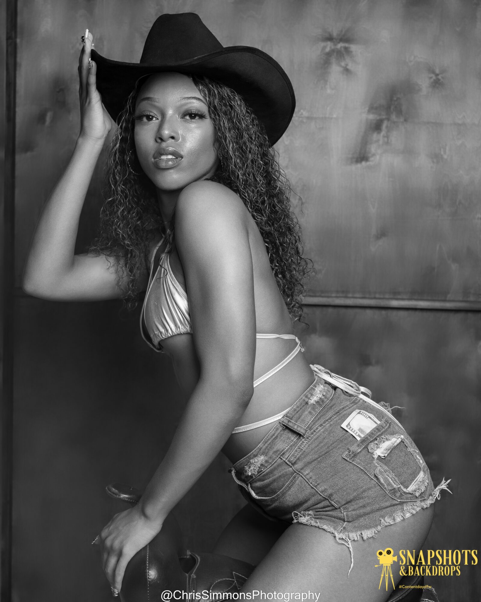 Young woman wearing a hat and bikini top, posing against a textured wall, with one hand on her head and the other on her hip.