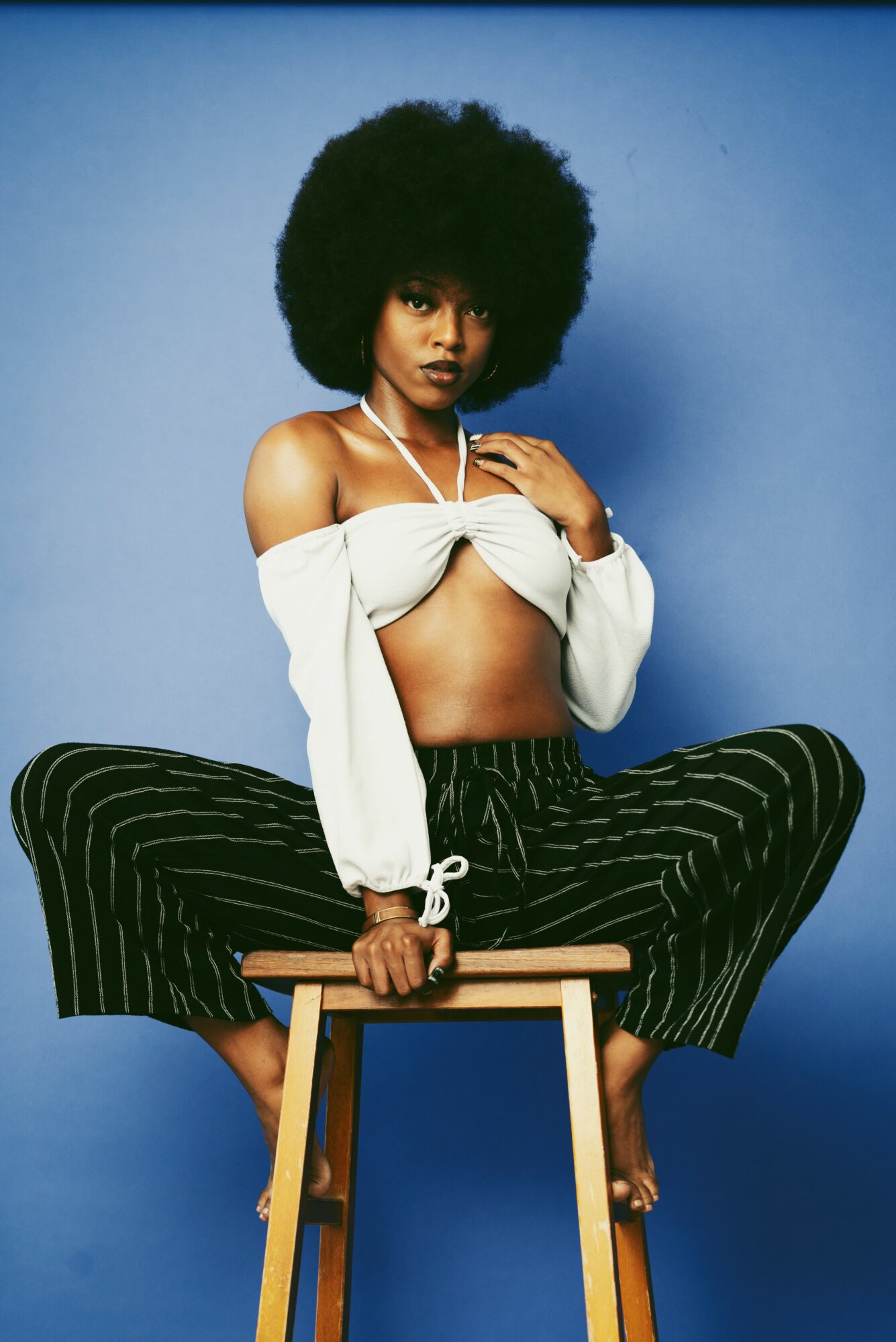 Young woman with large afro hairstyle sitting on a stool against a blue background, wearing a white off-shoulder crop top and striped pants.