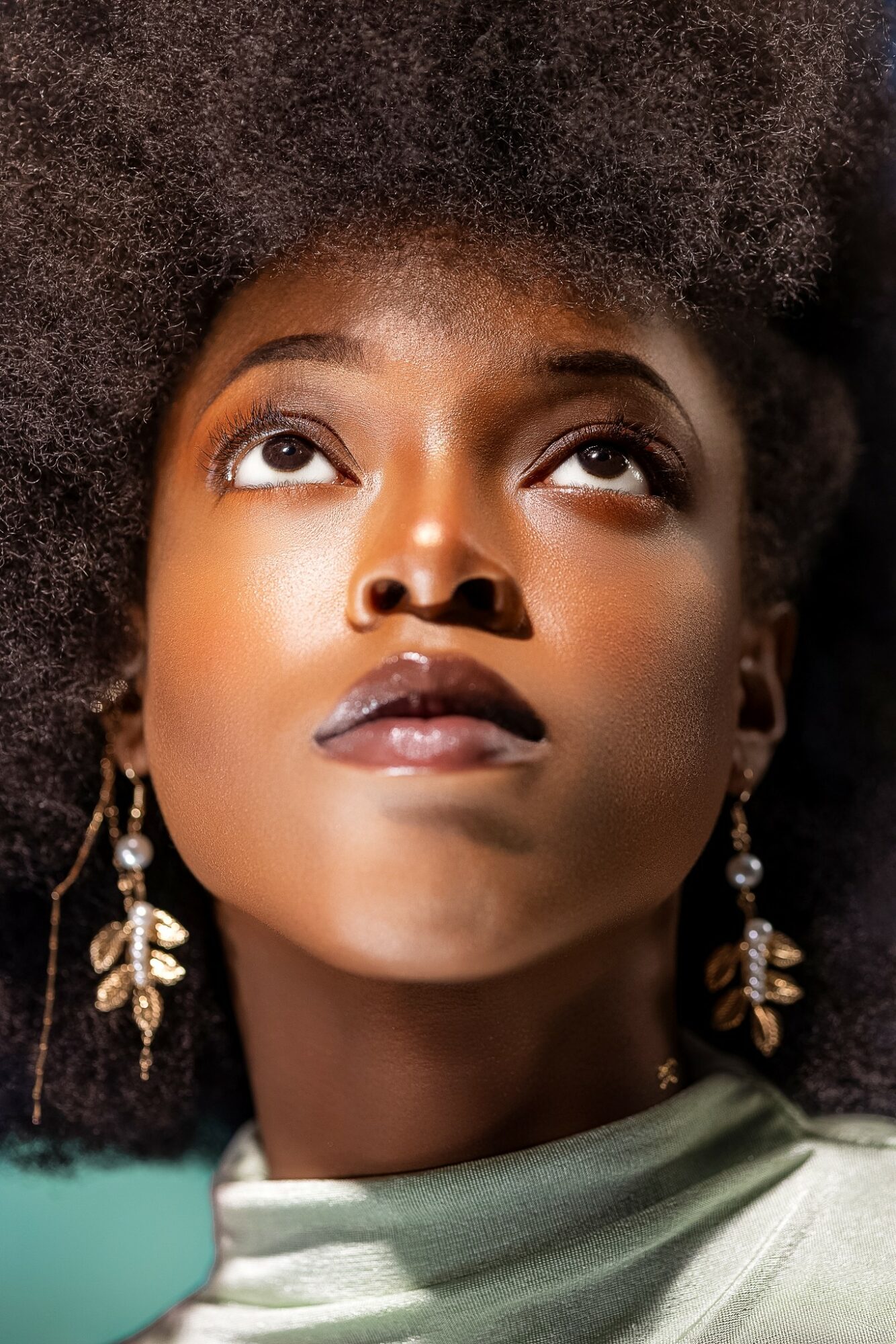 Close-up of a woman with natural hair looking upward, wearing earrings and a light-colored top.