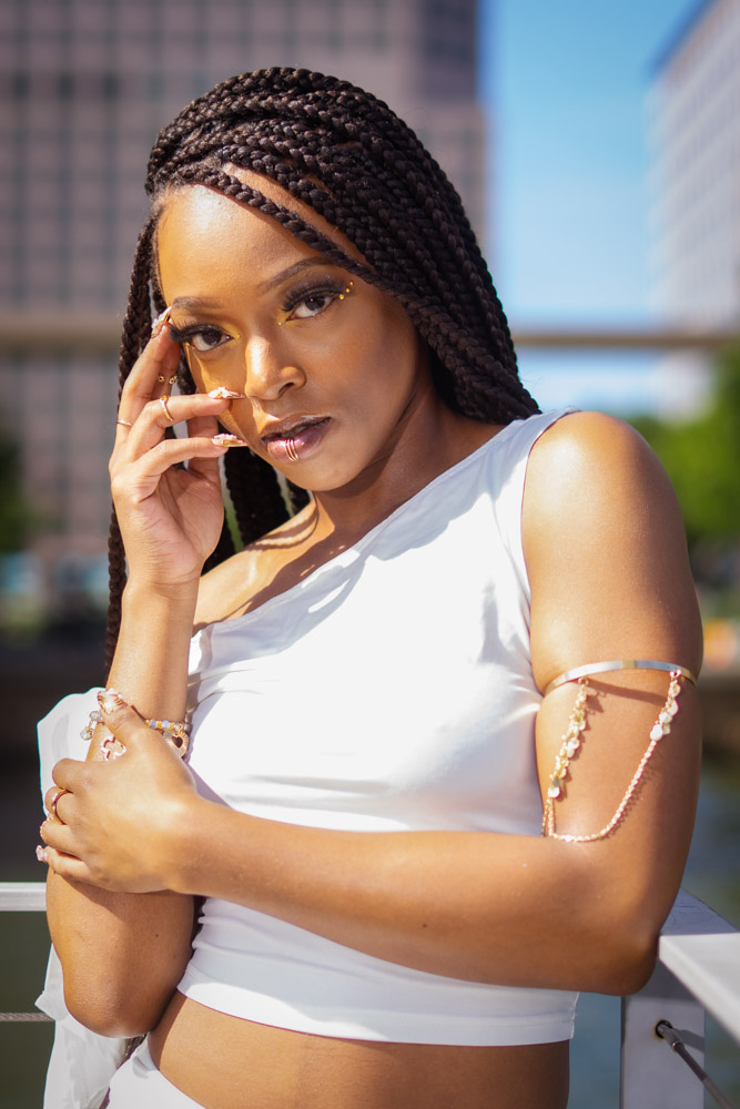 Young woman with braided hair wearing a white sleeveless top outdoors, with buildings and blue sky in background.