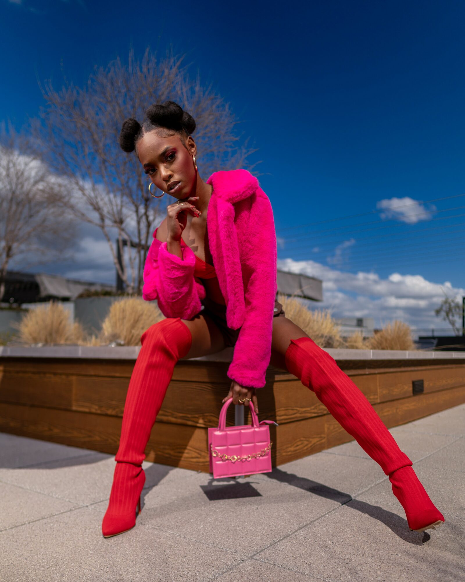 Young woman in bright pink outfit sitting outdoors on a wooden bench, holding a matching pink handbag, with a clear blue sky.
