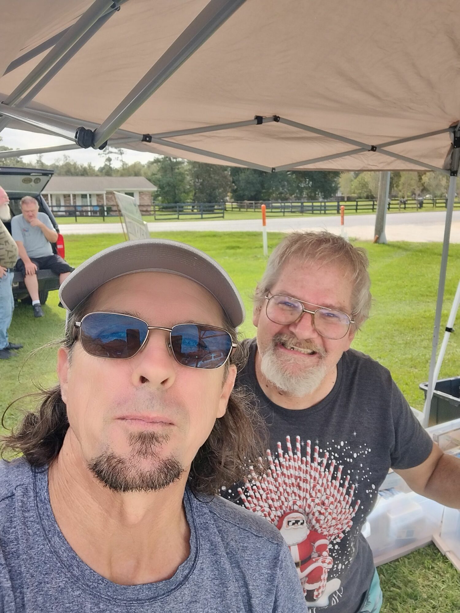Two men outdoors under a canopy, one wearing sunglasses and a cap, the other with glasses, both smiling.