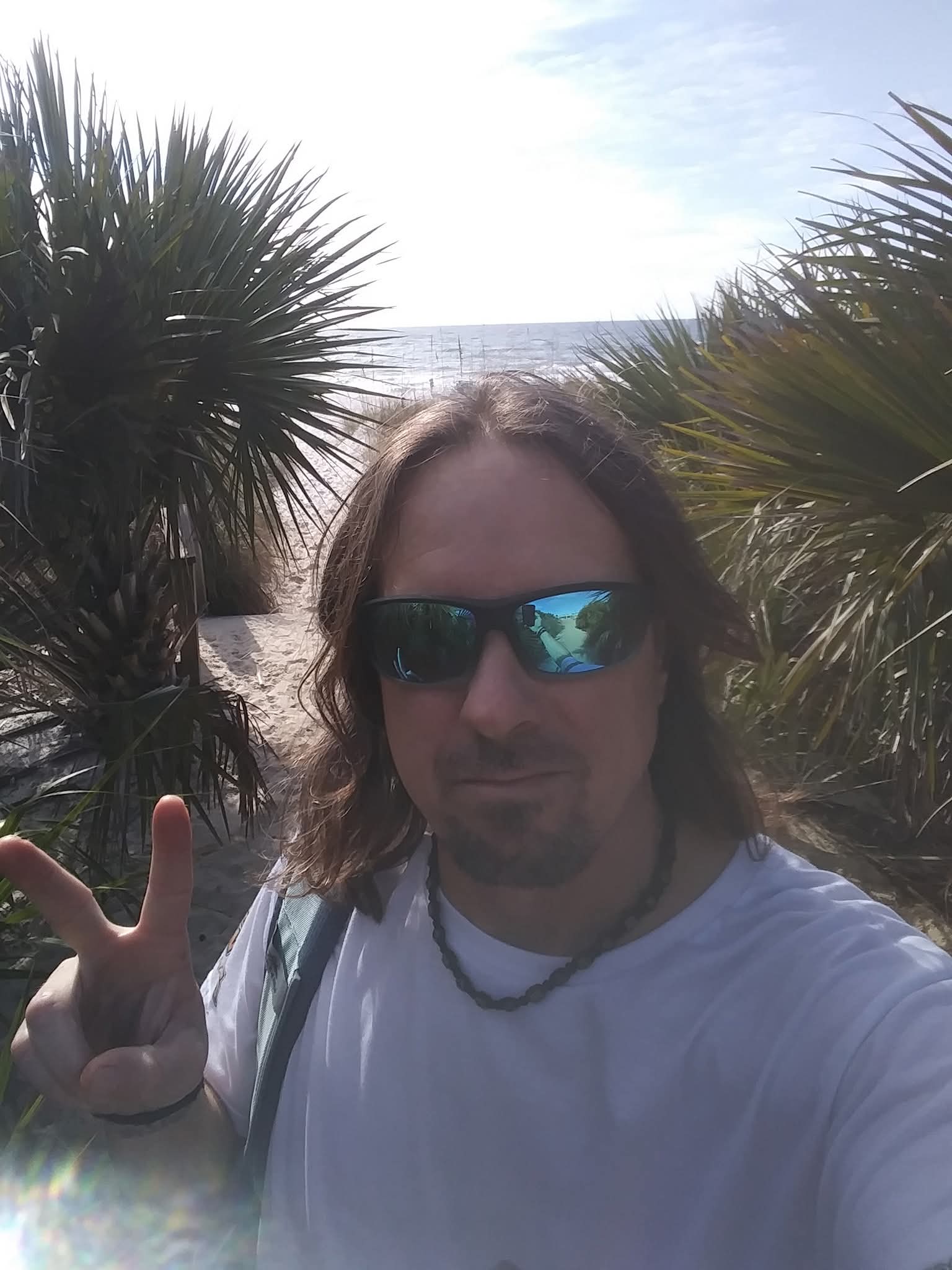 Person with long hair wearing sunglasses and a white shirt, making a peace sign outdoors near palm trees.