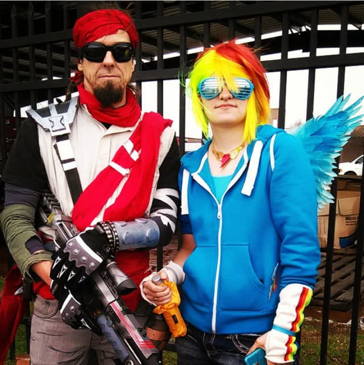 Two people in colorful costumes stand outdoors behind a black fence, one with rainbow hair and wings, the other with red hair and sunglasses.