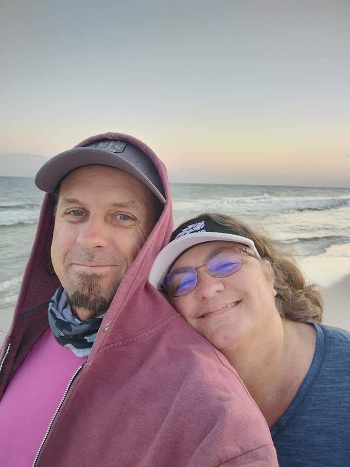 Two people smiling at the beach, one wearing a cap and the other with glasses, close together with ocean in background.