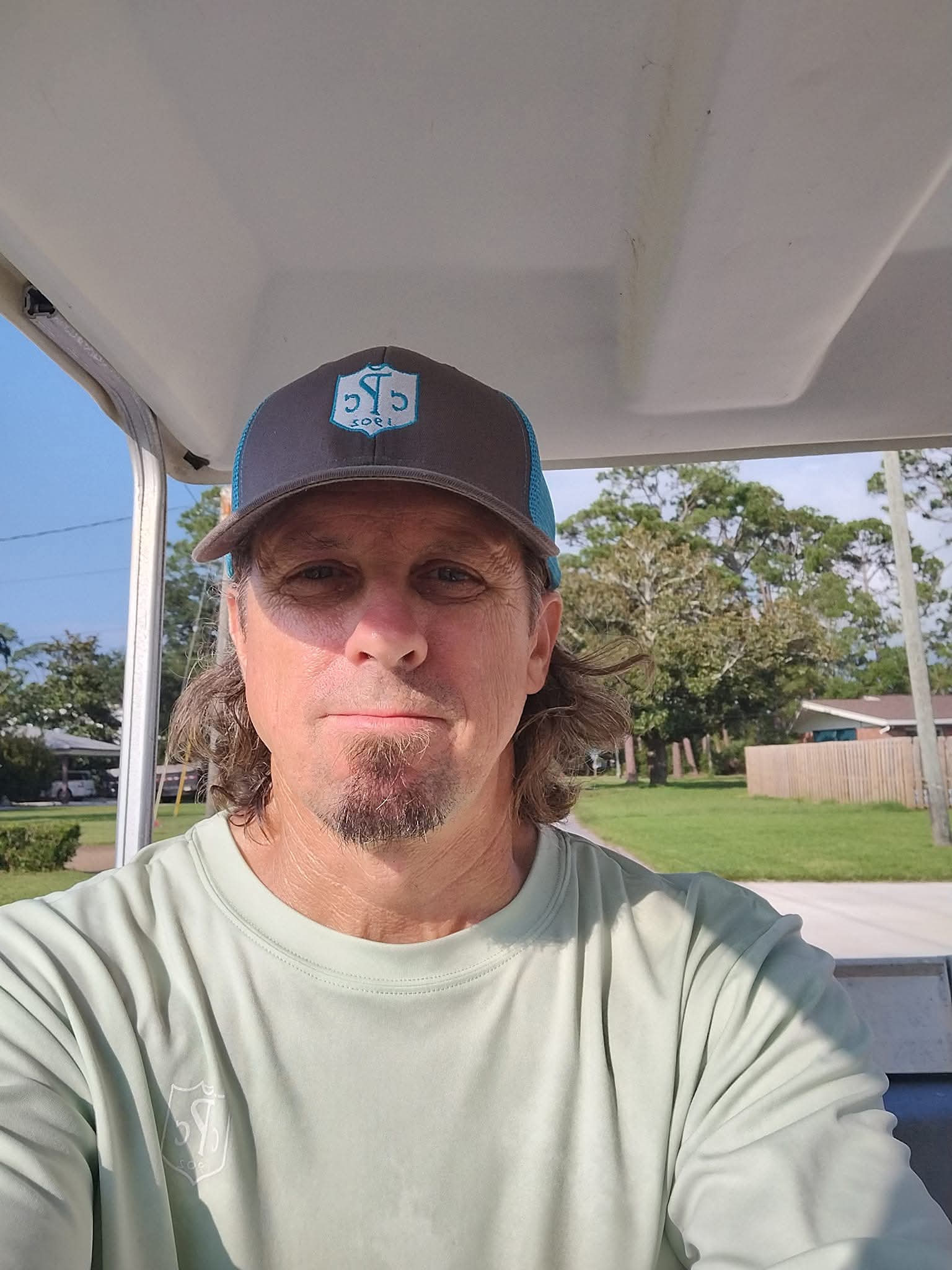 Man with long hair and beard wearing a baseball cap takes selfie outdoors on a sunny day.