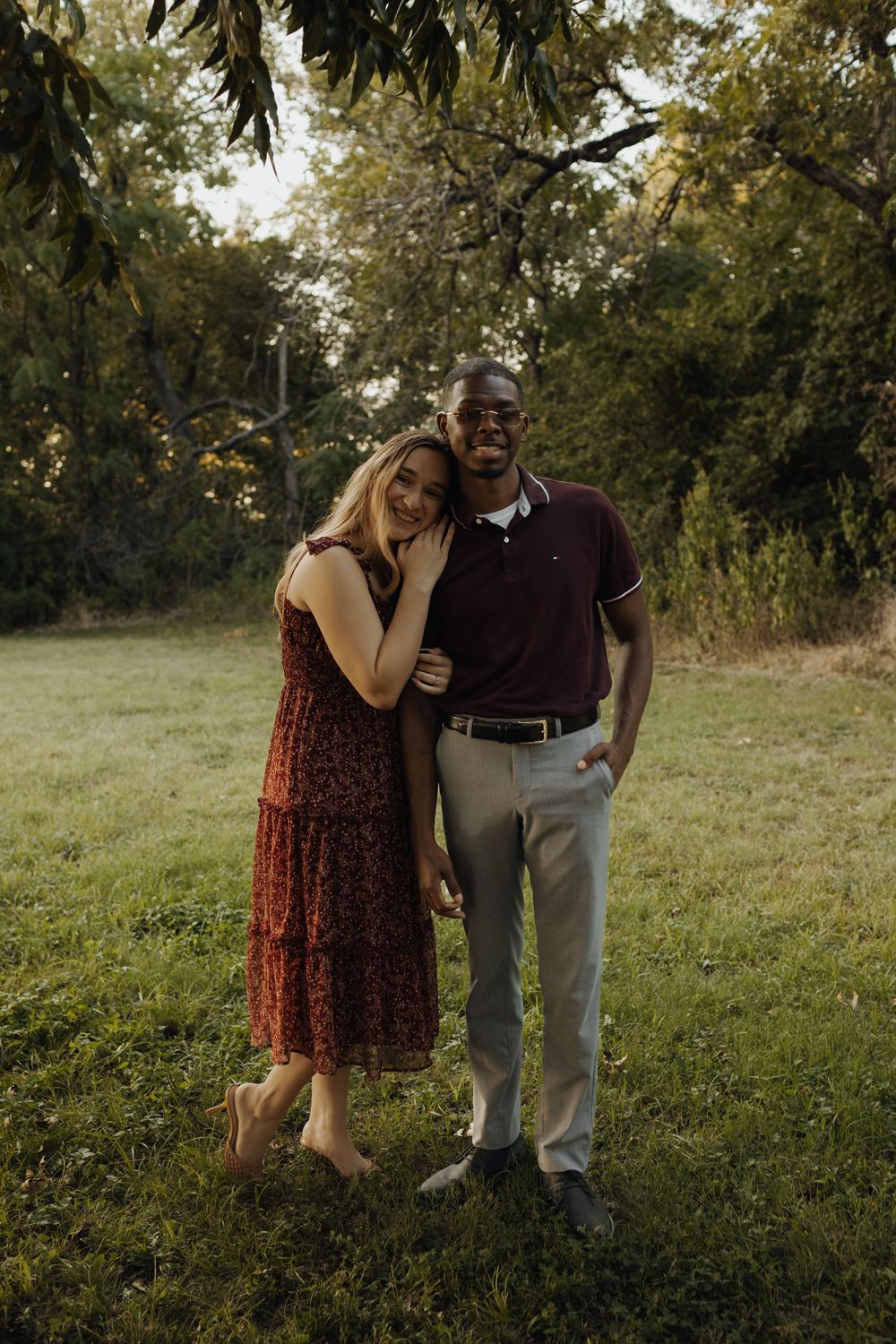 A woman and a man stand together outdoors, surrounded by trees and grass, smiling at the camera.