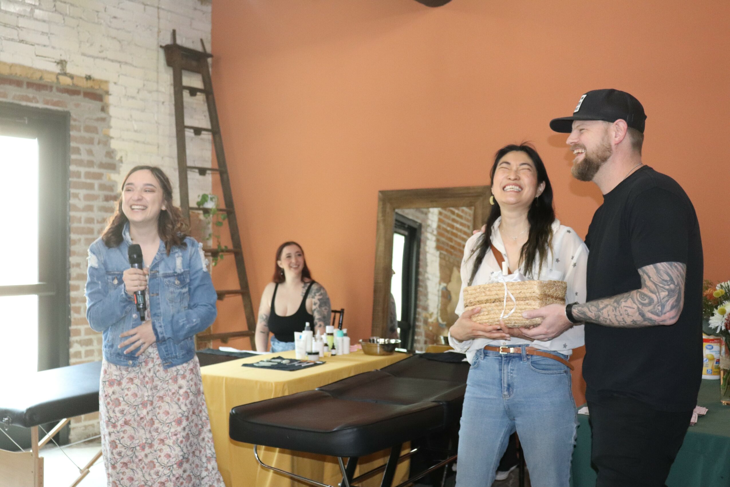 Group of four people in a room, one woman holding a microphone, others smiling and talking, with a ladder and window in the background.