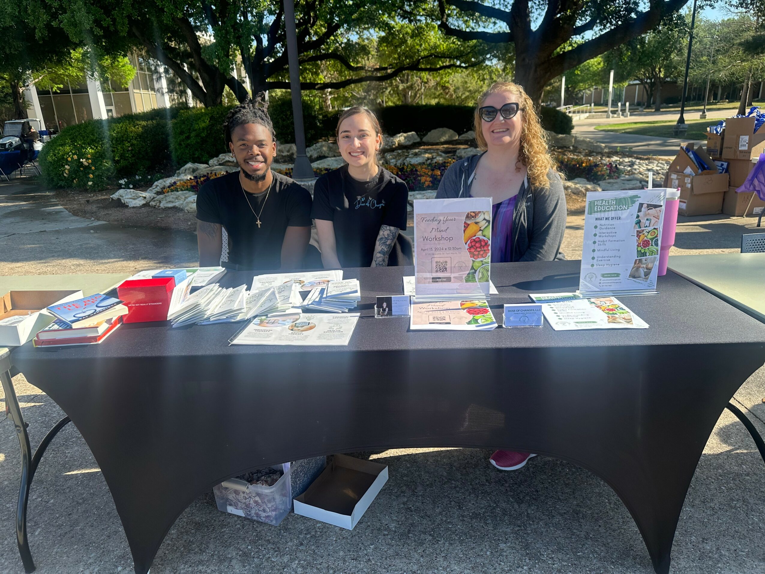 Three women sit behind a table outdoors with informational displays and books, trees and a park in the background.