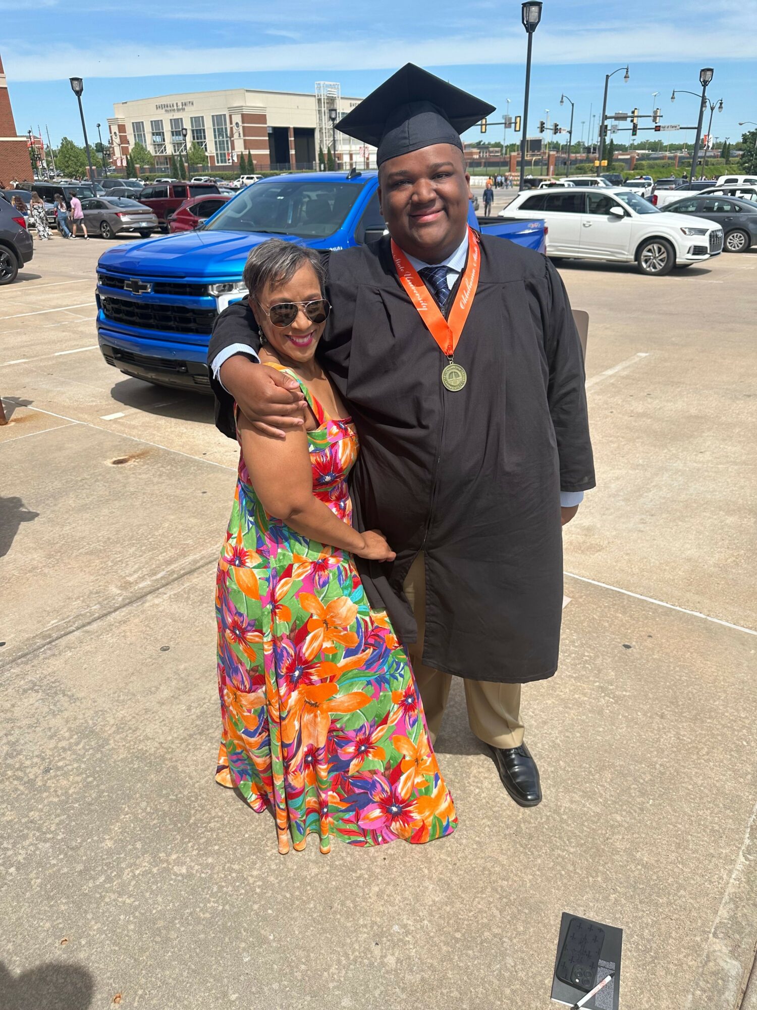 Person in graduation gown and cap standing next to a woman in colorful dress, both smiling, in a parking lot.