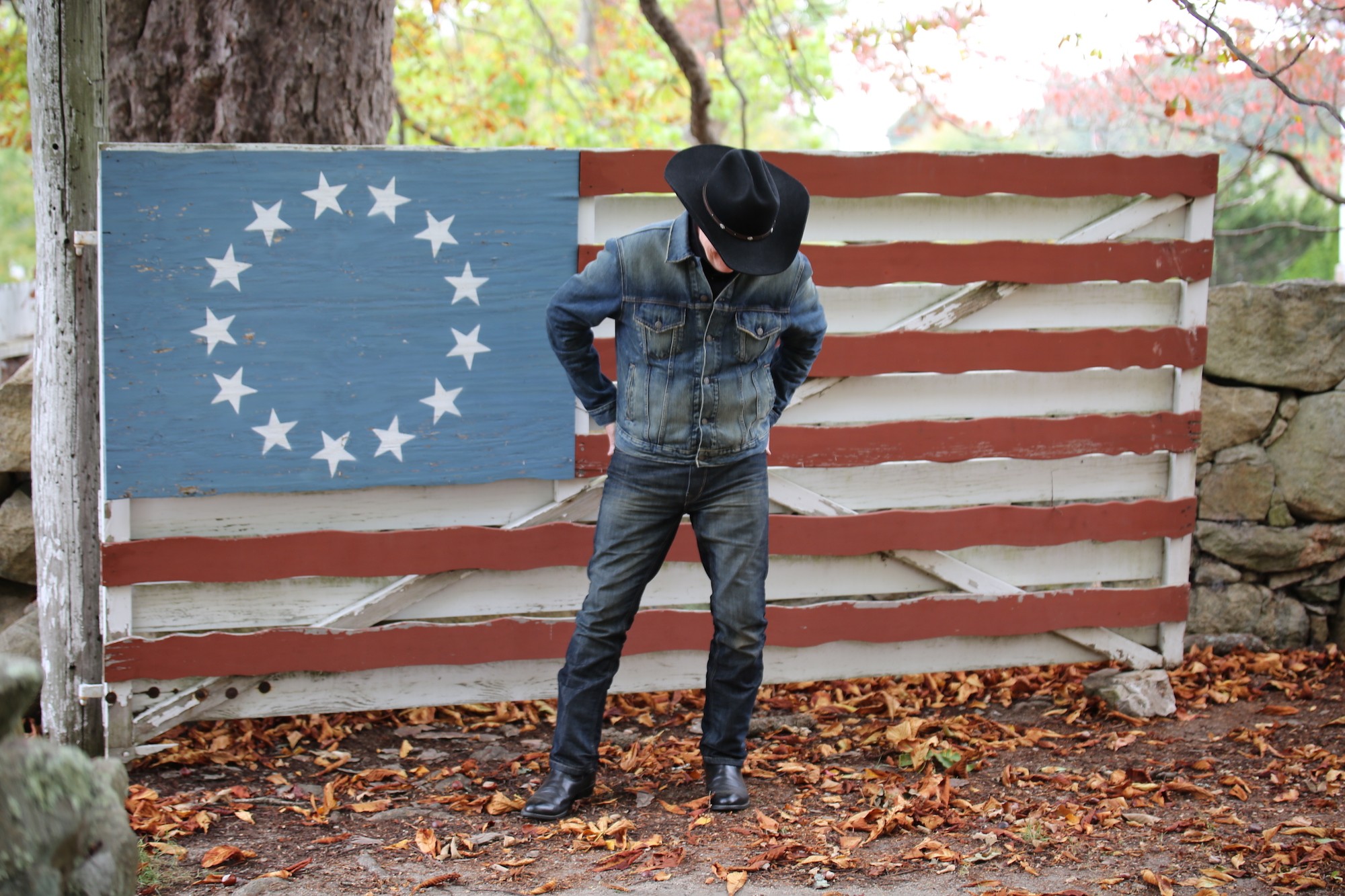 Person standing in front of a painted American flag with a blue circle and white stars, outdoors with fallen leaves.