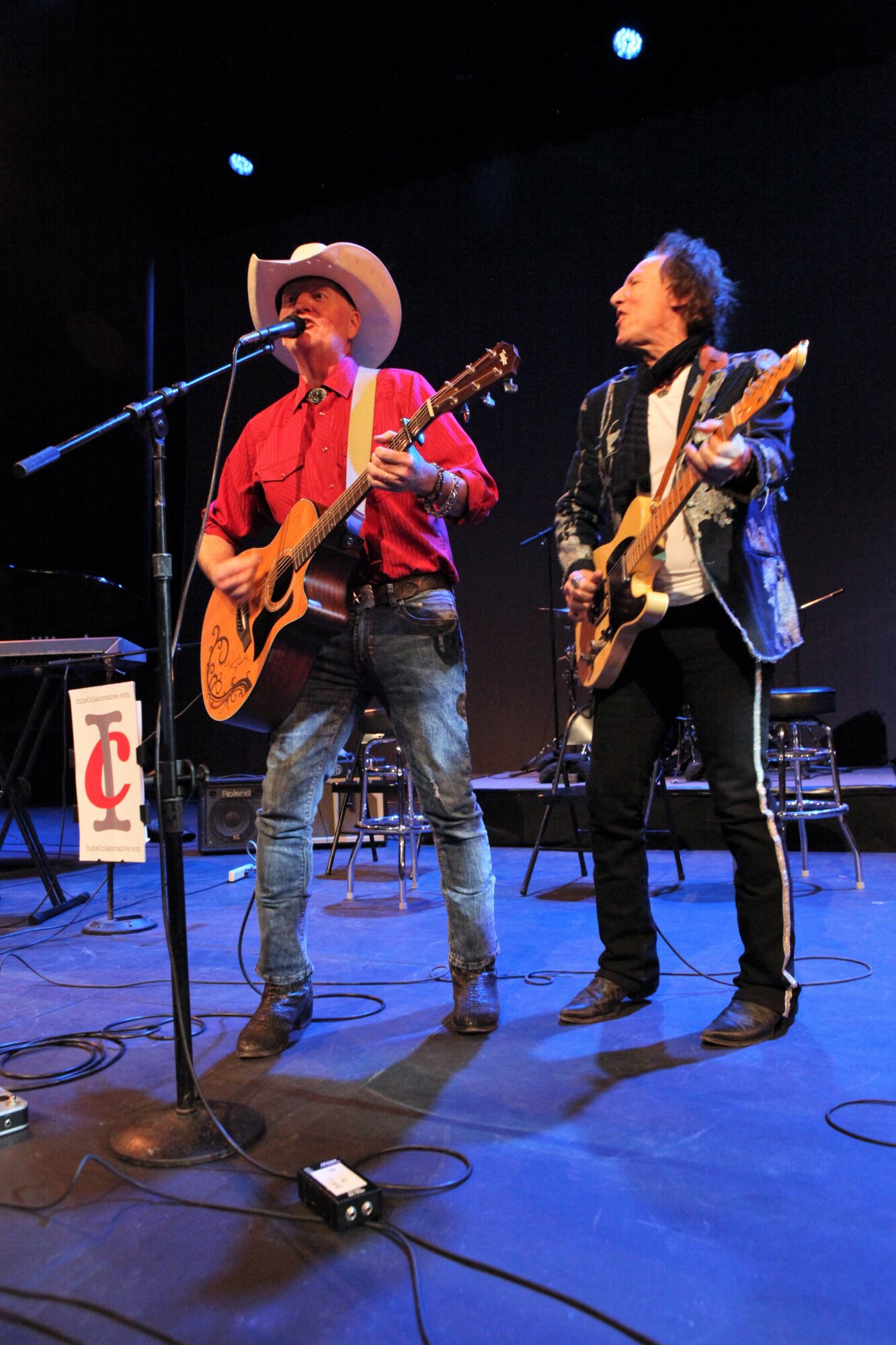 Two musicians perform on stage with guitars, one wearing a cowboy hat and red shirt, the other in a shiny jacket.