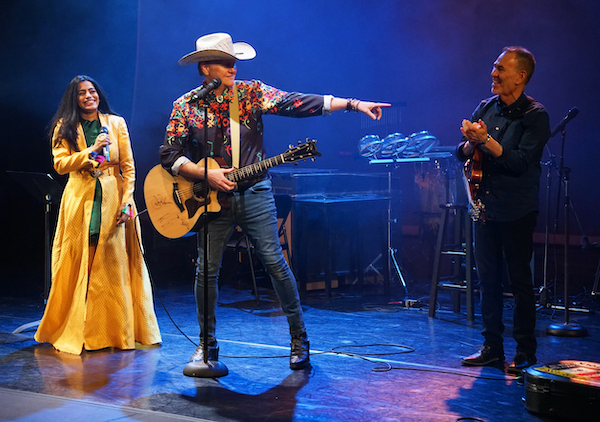 Three performers on stage: a woman in a yellow dress, a man with a guitar and cowboy hat, and another man clapping.