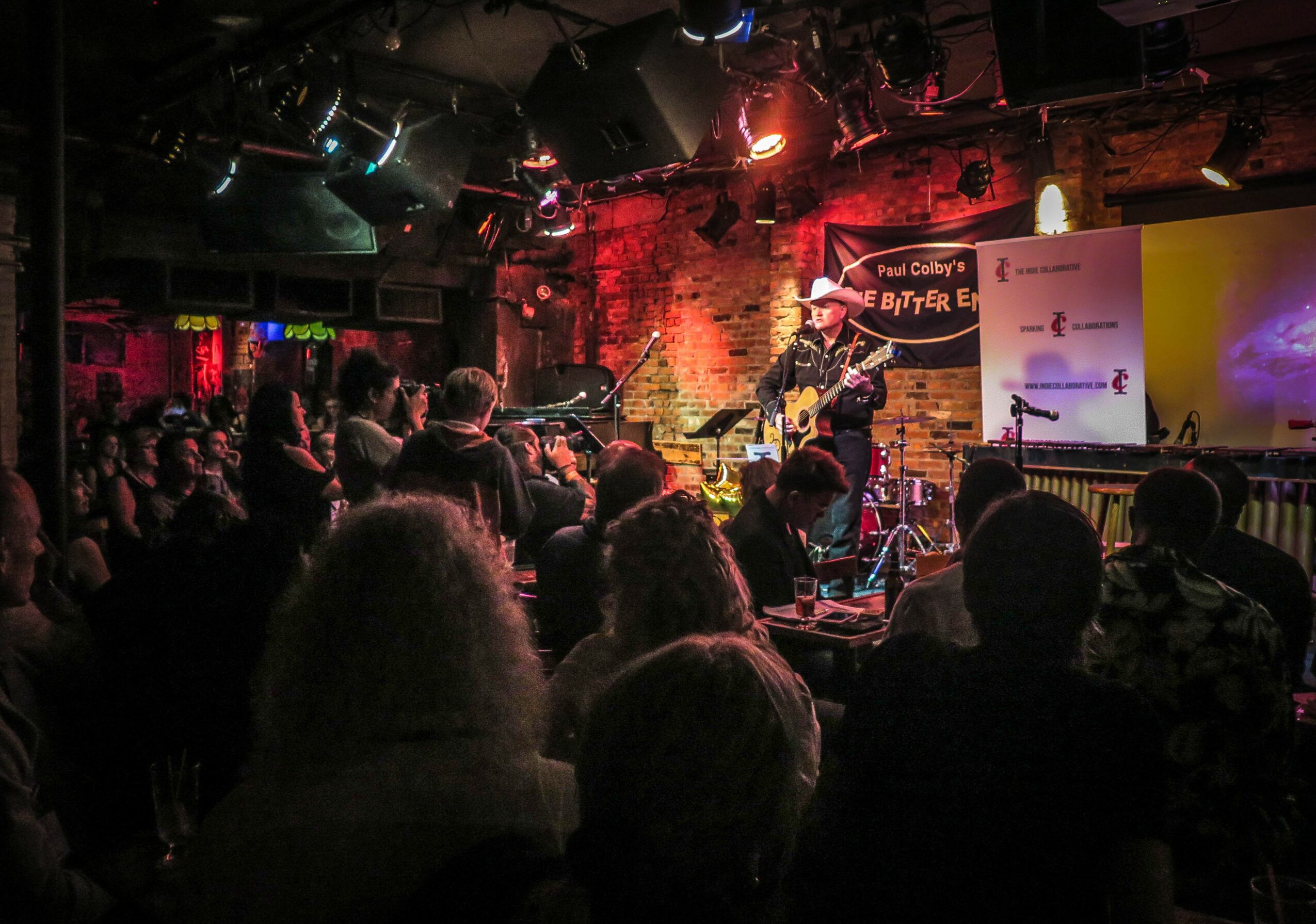 Performer on stage with guitar in a dimly lit venue, audience watching attentively, brick wall background, colorful lights.