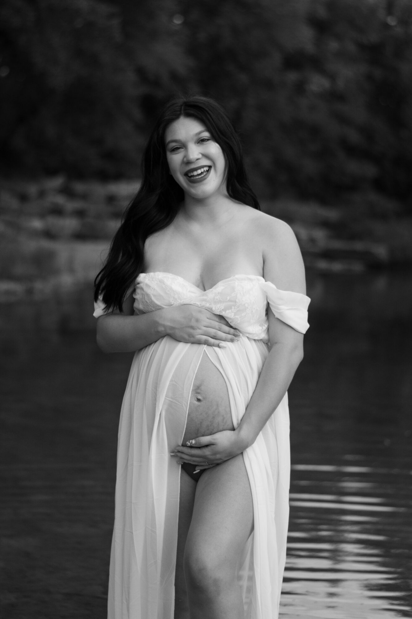 Pregnant woman in a flowing dress standing outdoors near water, smiling, with trees in the background.