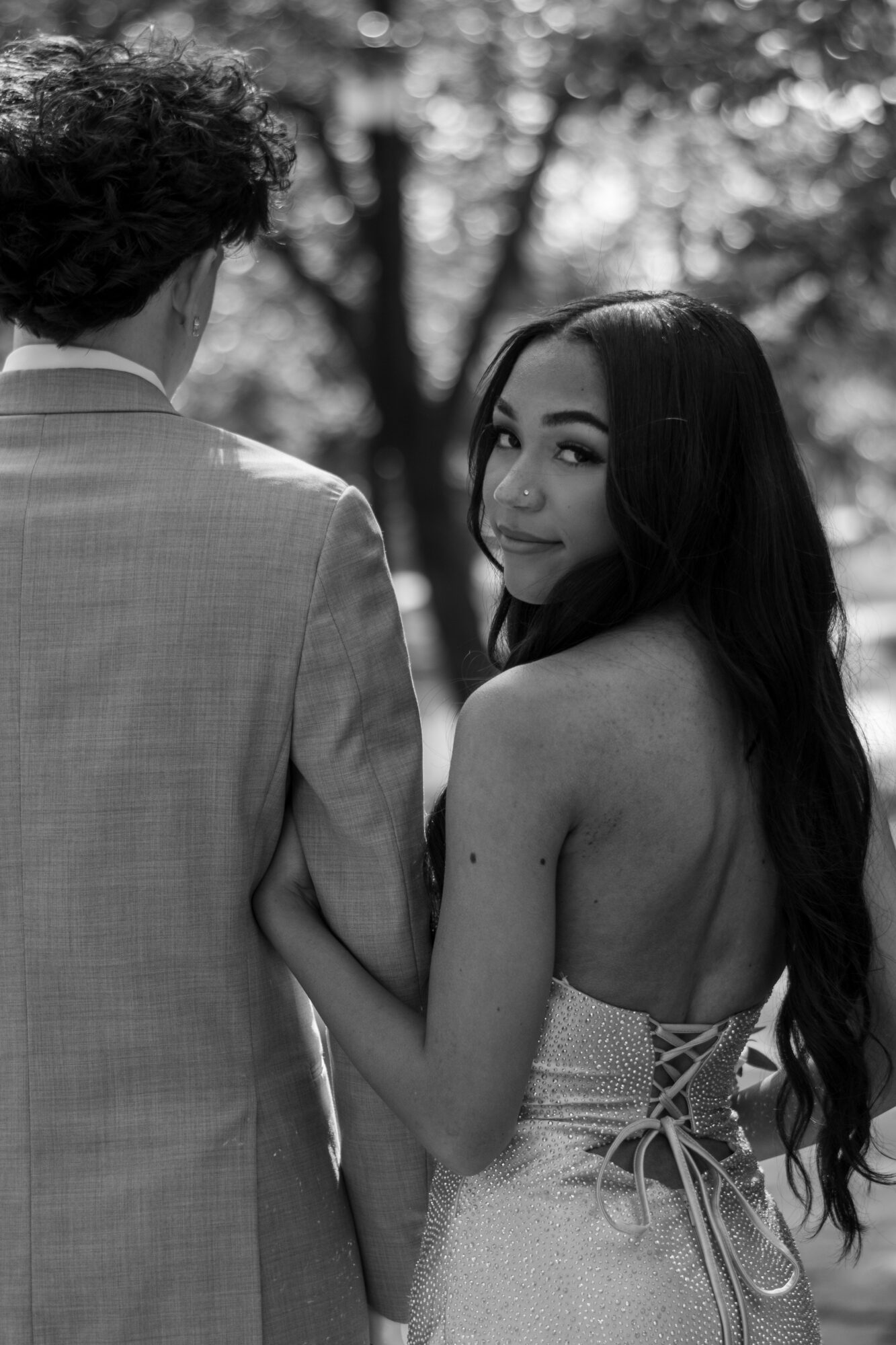 A woman with long dark hair and a strapless dress looks over her shoulder at a man in a suit outdoors.