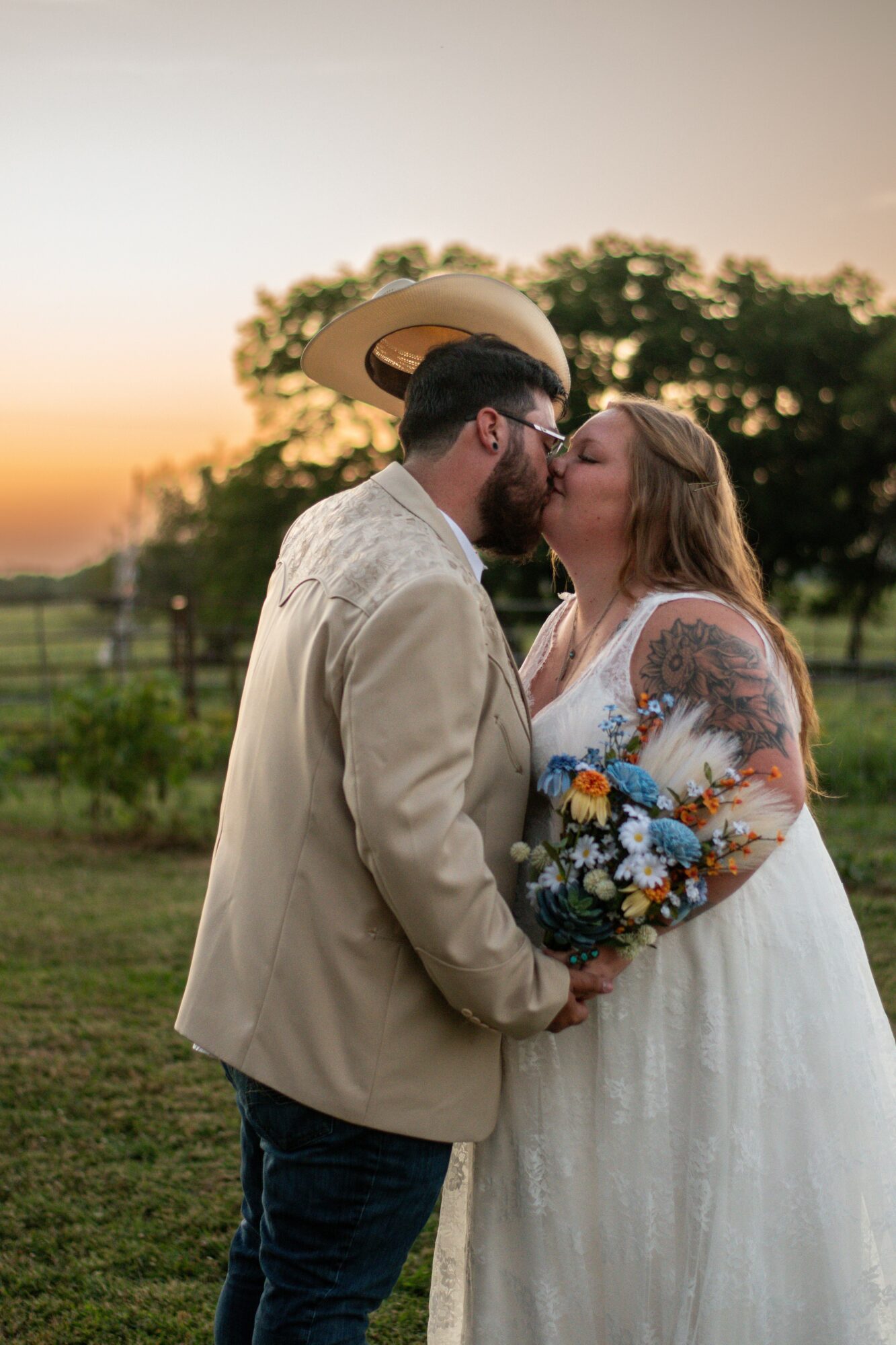 Couple kissing outdoors at sunset, woman holding a bouquet, man wearing a hat, trees in background.