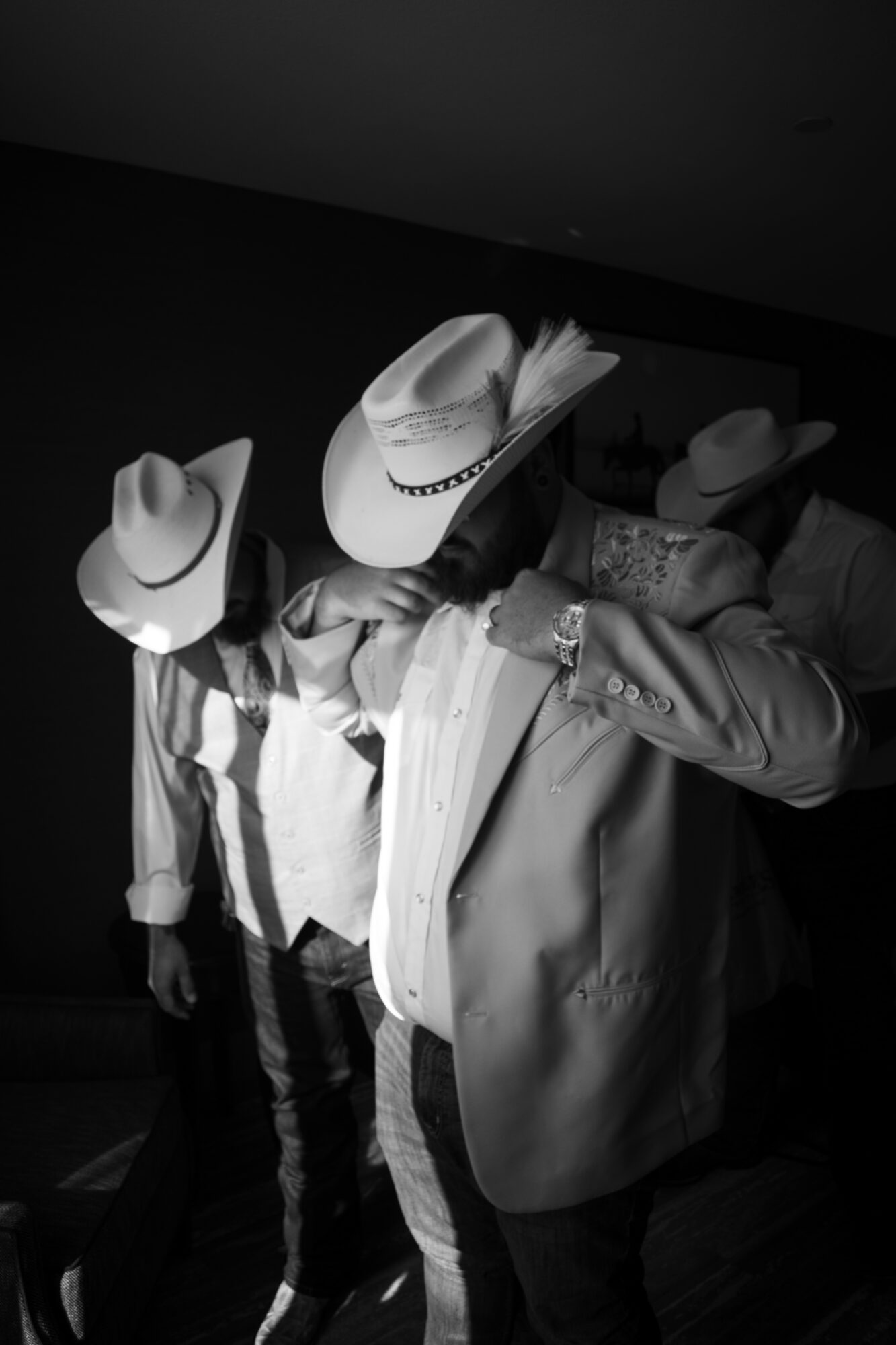Group of people wearing cowboy hats, some adjusting hats, in a dark indoor setting, black and white photo.