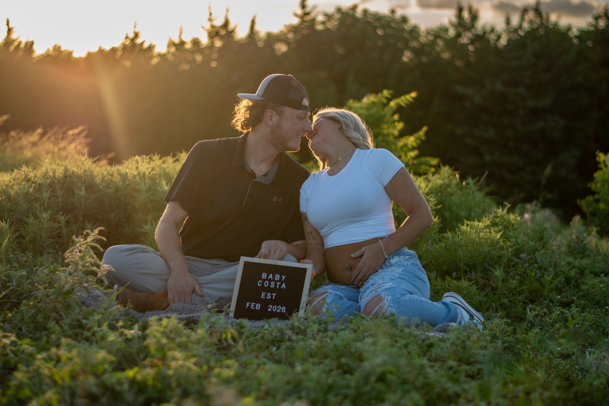 Pregnant woman and man sitting on grass, touching foreheads, with a sign reading 'Baby Due Est June 2023' in front, outdoors during sunset.