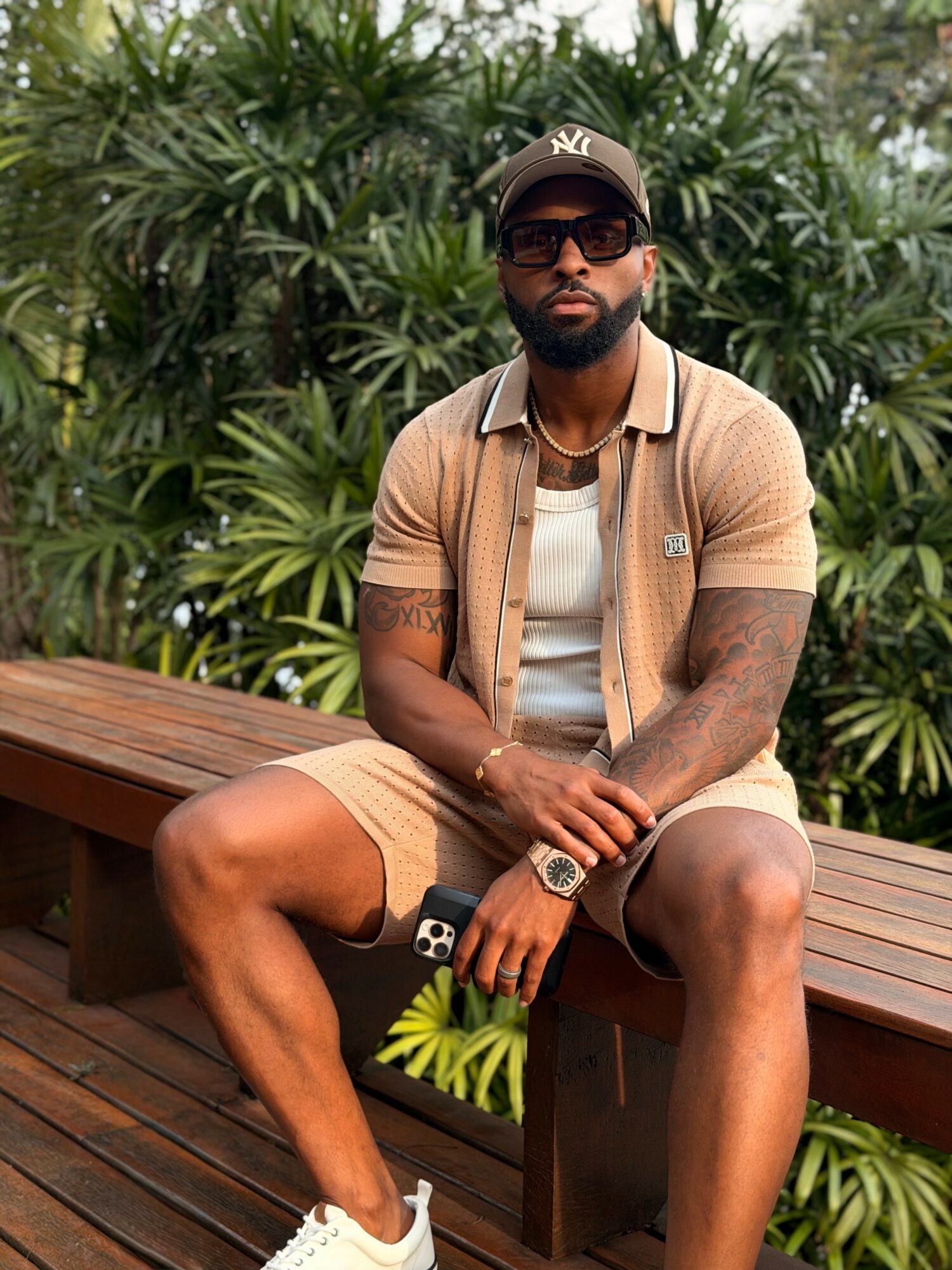 Man sitting on a wooden bench outdoors, wearing sunglasses, a cap, and casual clothing, with lush green plants in background.