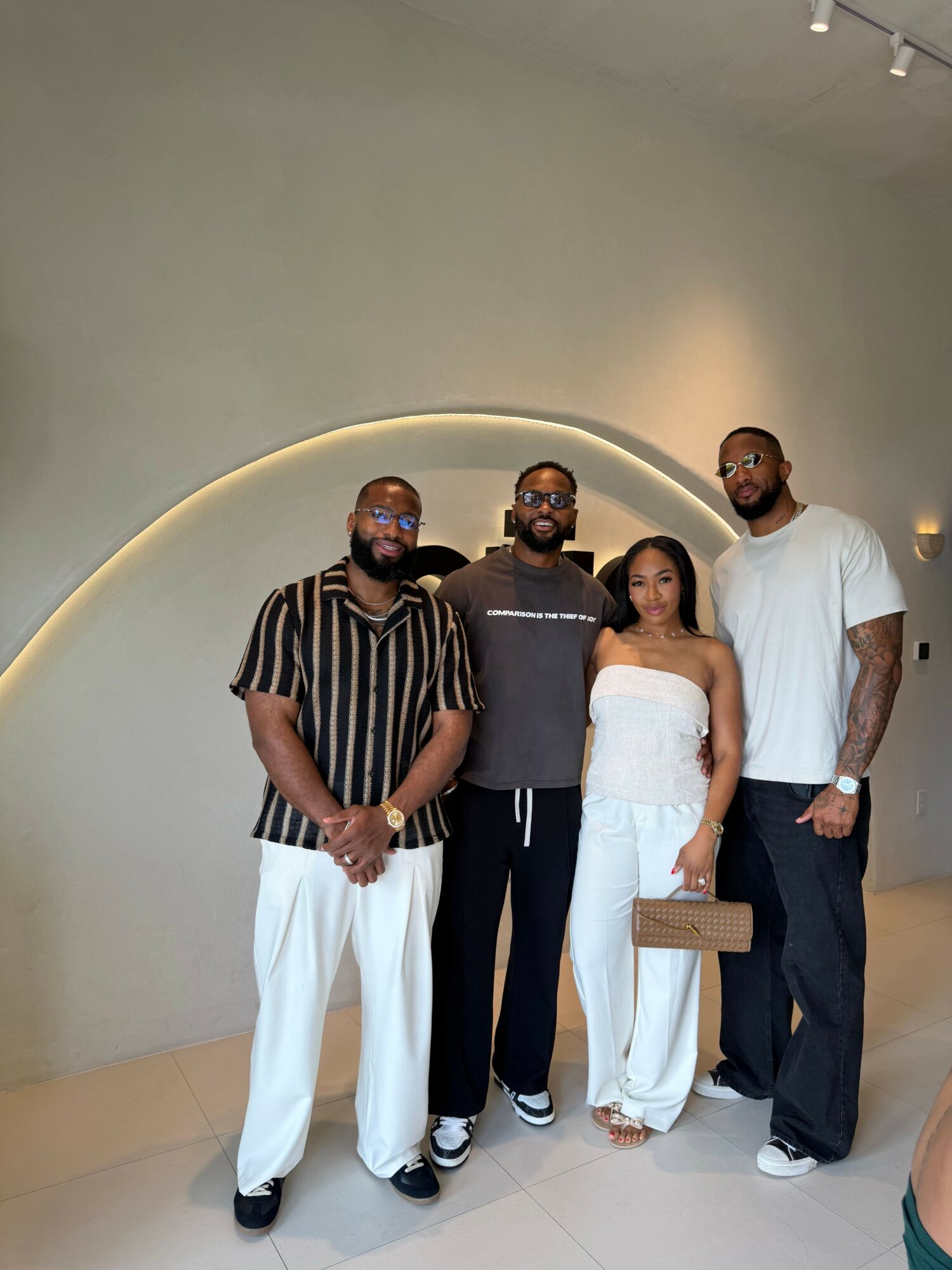 Group of five people standing indoors, some holding a brown paper bag, with a curved wall and lighting behind them.