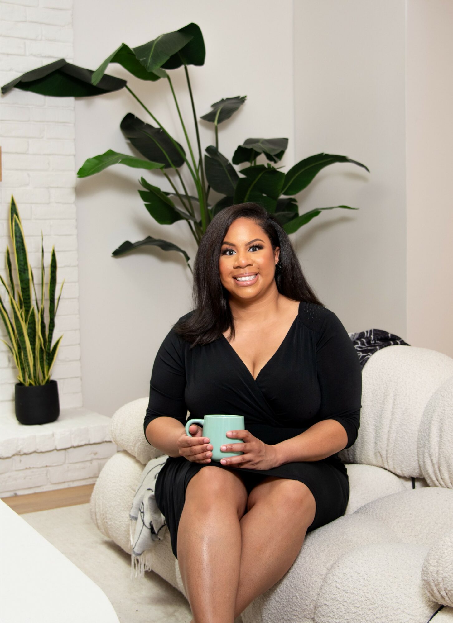 Woman sitting on a white sofa holding a mug, with large green plants behind her, in a bright room.