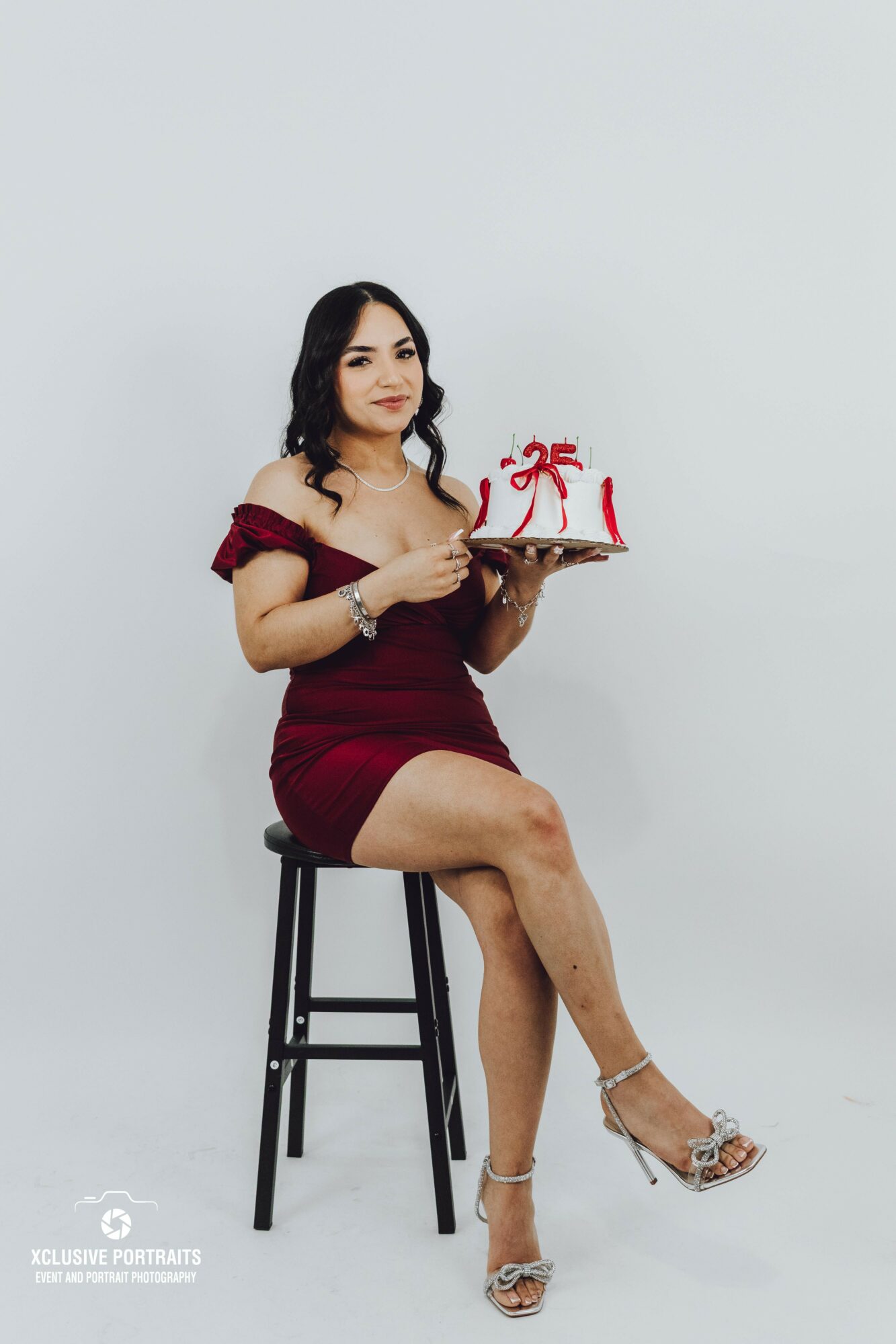 Woman in red dress sitting on a stool holding a cake with red ribbons, against a plain light background.