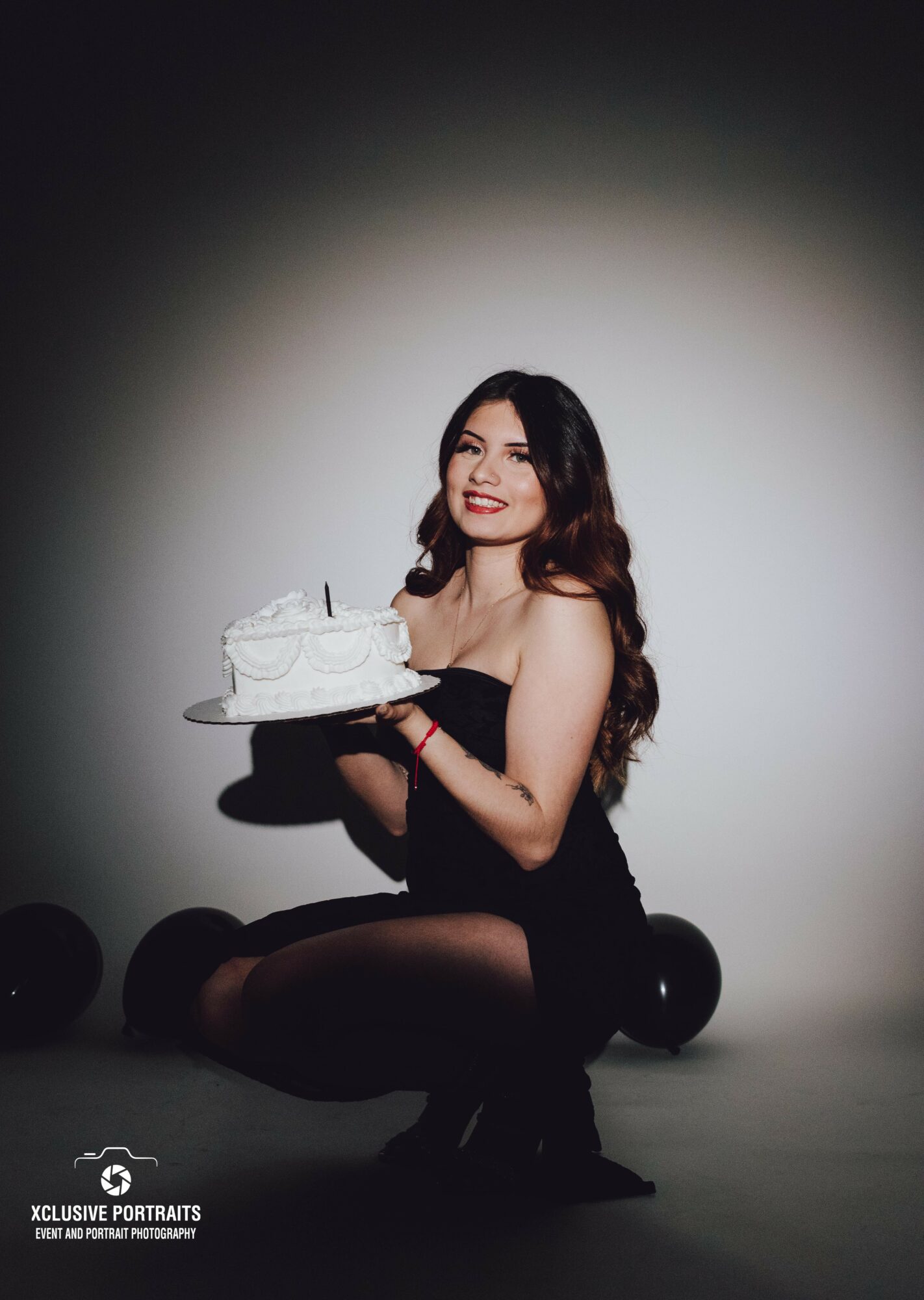 Woman with long dark hair in black dress holding a birthday cake, smiling, sitting on the floor against a plain background.
