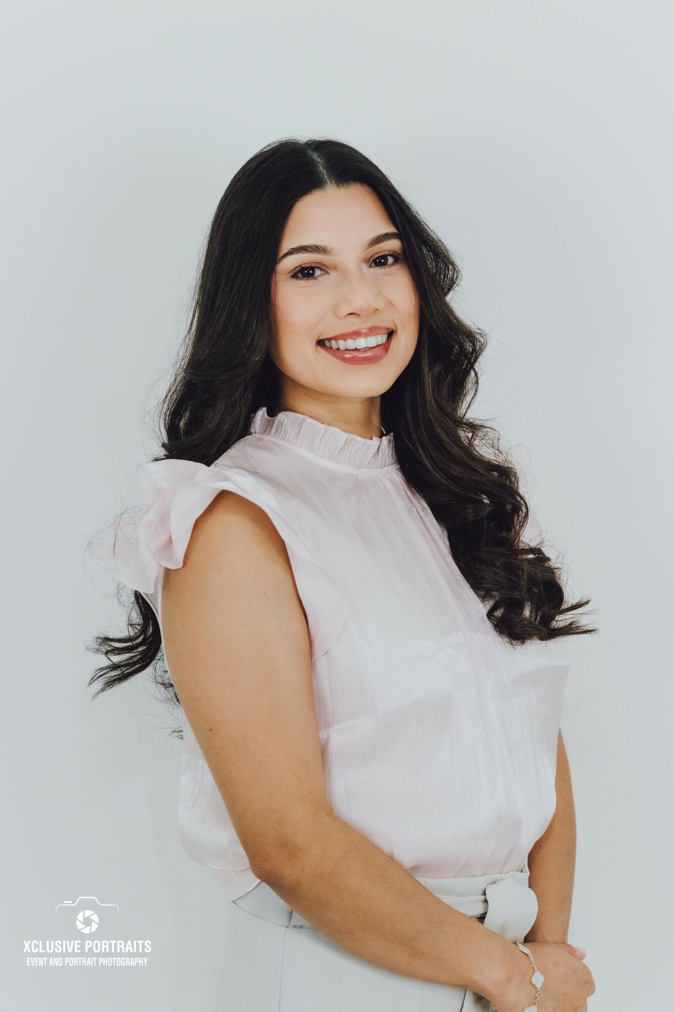 Young woman with long dark hair smiling, wearing a white sleeveless top, standing against a light background.
