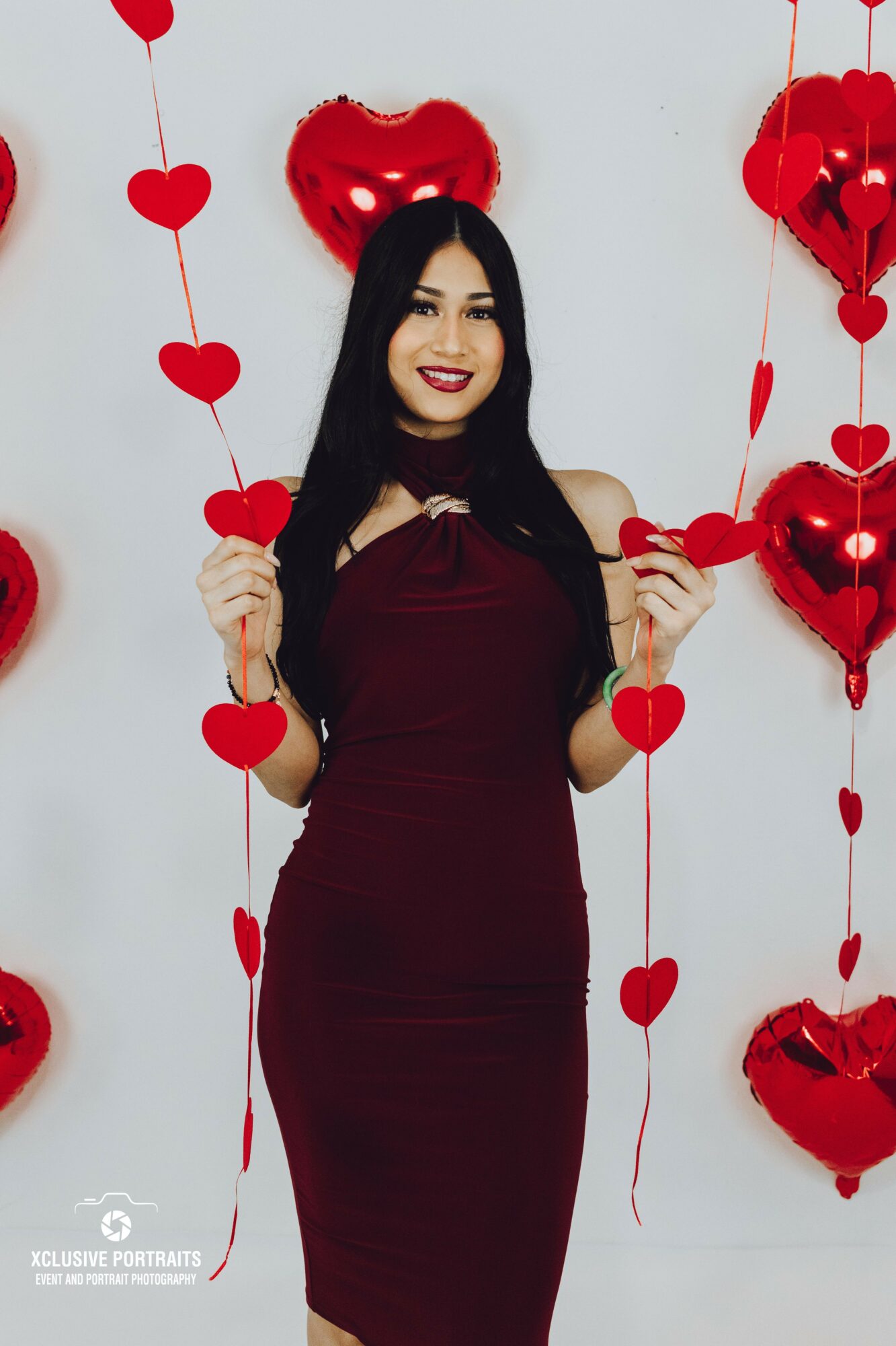 Woman in a burgundy dress holding heart-shaped decorations, standing in front of a wall with red heart balloons, smiling.