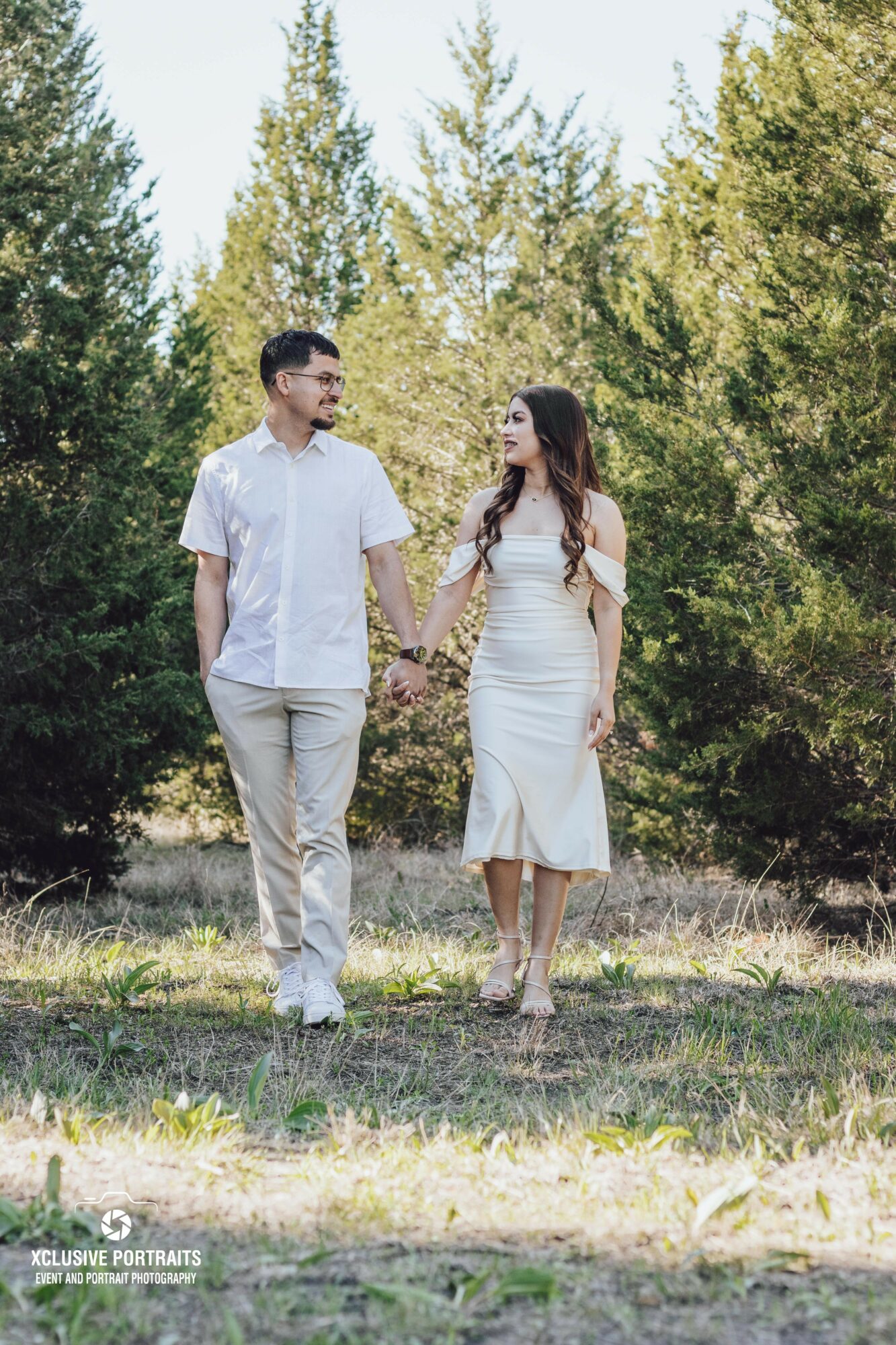 A couple holding hands walking outdoors in a grassy area with trees in the background, both smiling and looking at each other.