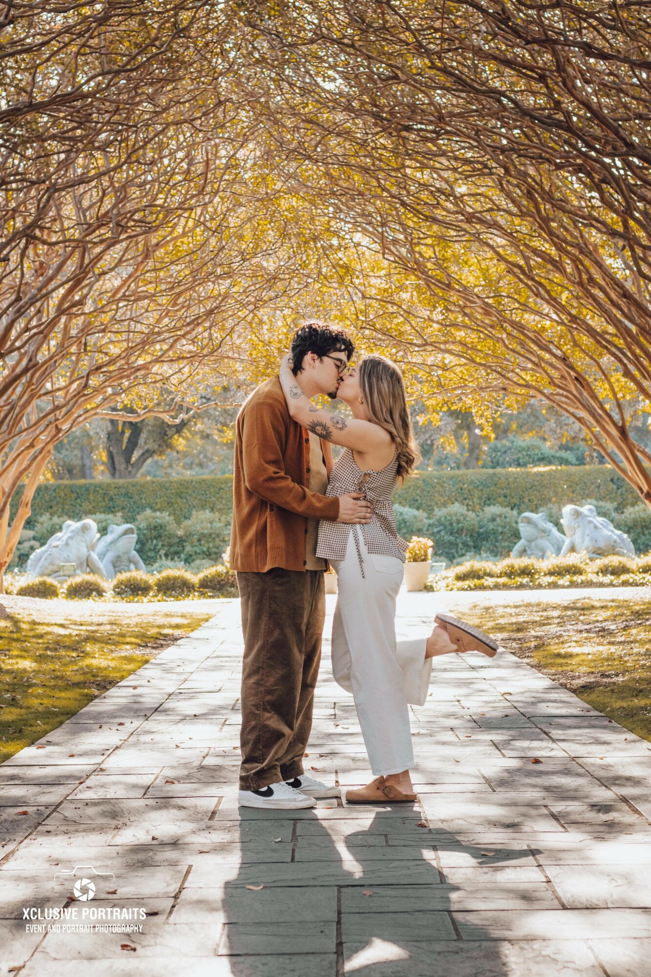 A couple embraces in a park with trees and fountains, sunlight filtering through branches, casting shadows on the ground.