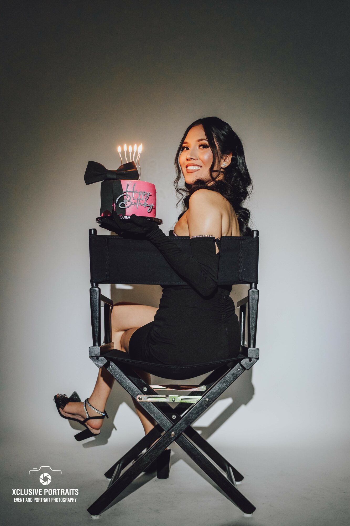 Woman sitting on a director's chair holding a birthday cake with lit candles, smiling, wearing a black dress, against a plain background.