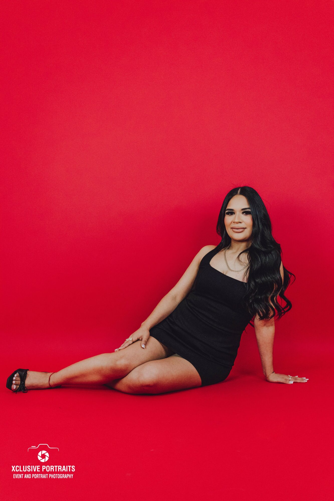 Woman in black dress sitting on red background, smiling, long dark hair, one leg extended, one arm supporting her, black heels